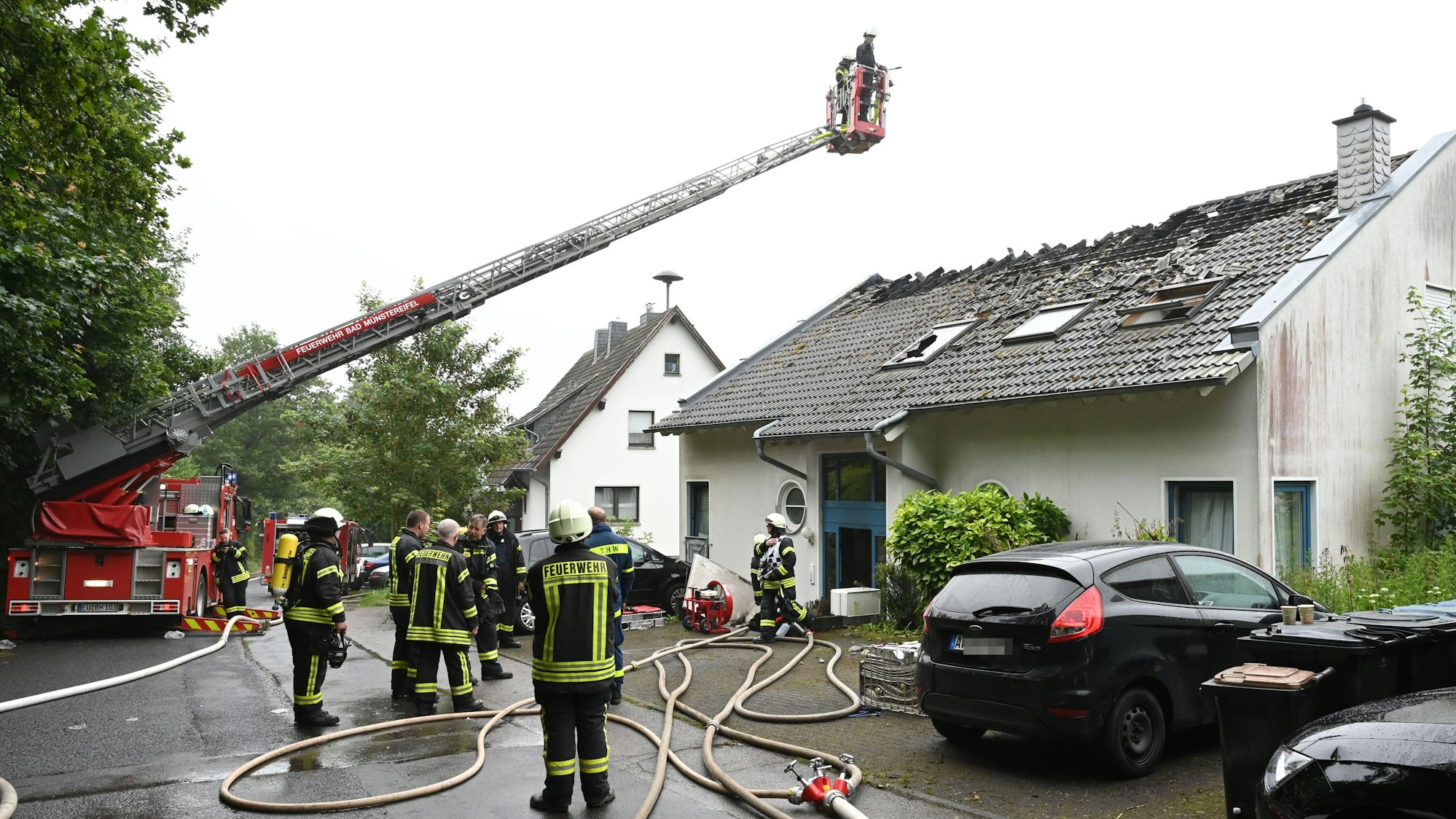Feuerwehrleute stehen vor dem Haus, in dem es gebrannt hat. Zwei Einsatzkräfte sind im Korb der Drehleiter und schauen auf das zerstörte Dach.