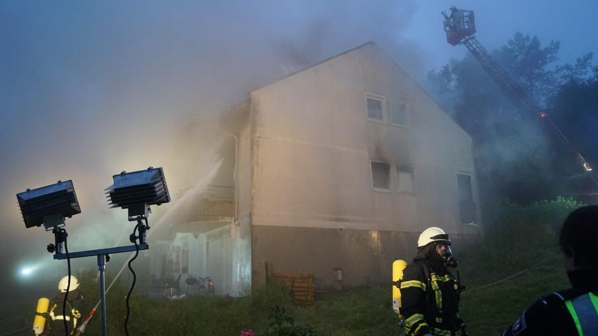 Das brennende Haus wird von zwei Strahlern angeleuchtet. Rauch ist in der Luft. Feuerwehrleute löschen den Brand.
