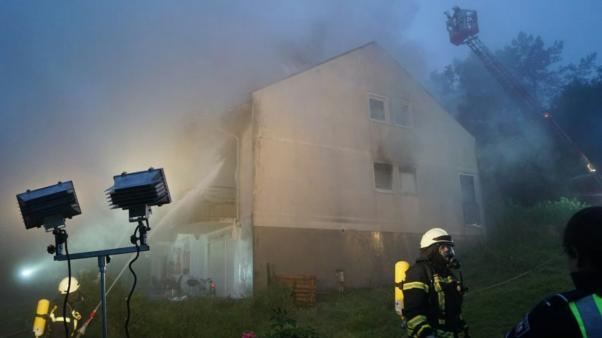 Das brennende Haus wird von zwei Strahlern angeleuchtet. Rauch ist in der Luft. Feuerwehrleute löschen den Brand.
