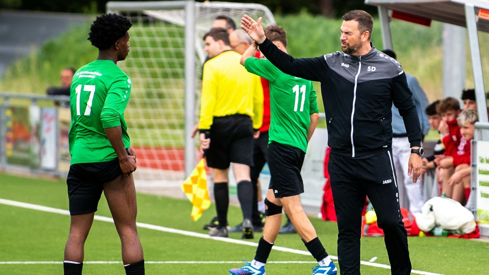 Ein Fußballtrainer gestikuliert mit einem Spieler, der zieht ein Hosenbein hoch.
