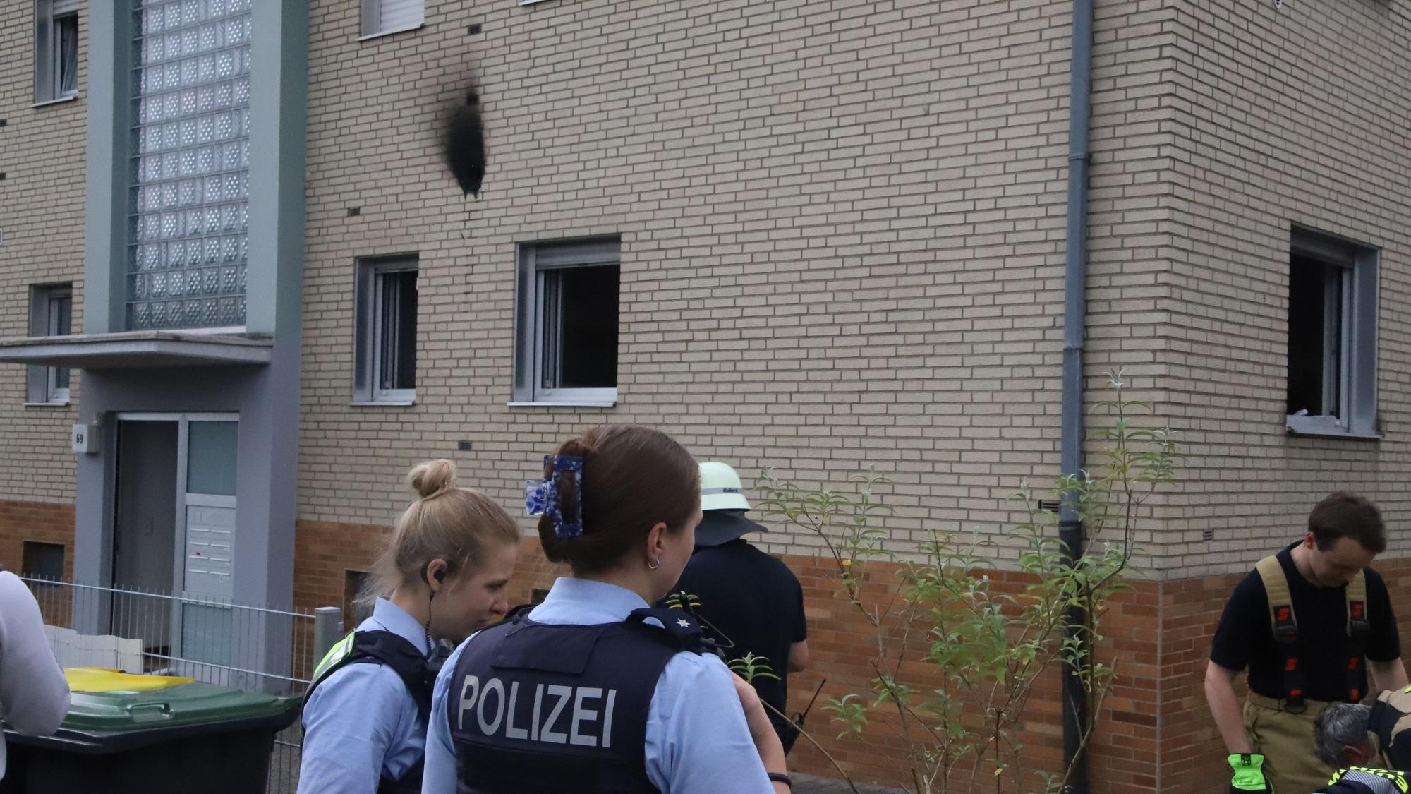 Polizistinnen und Feuerwehrleute stehen vor einem Haus, an dem ein schwarzer Rußfleck zu sehen ist.