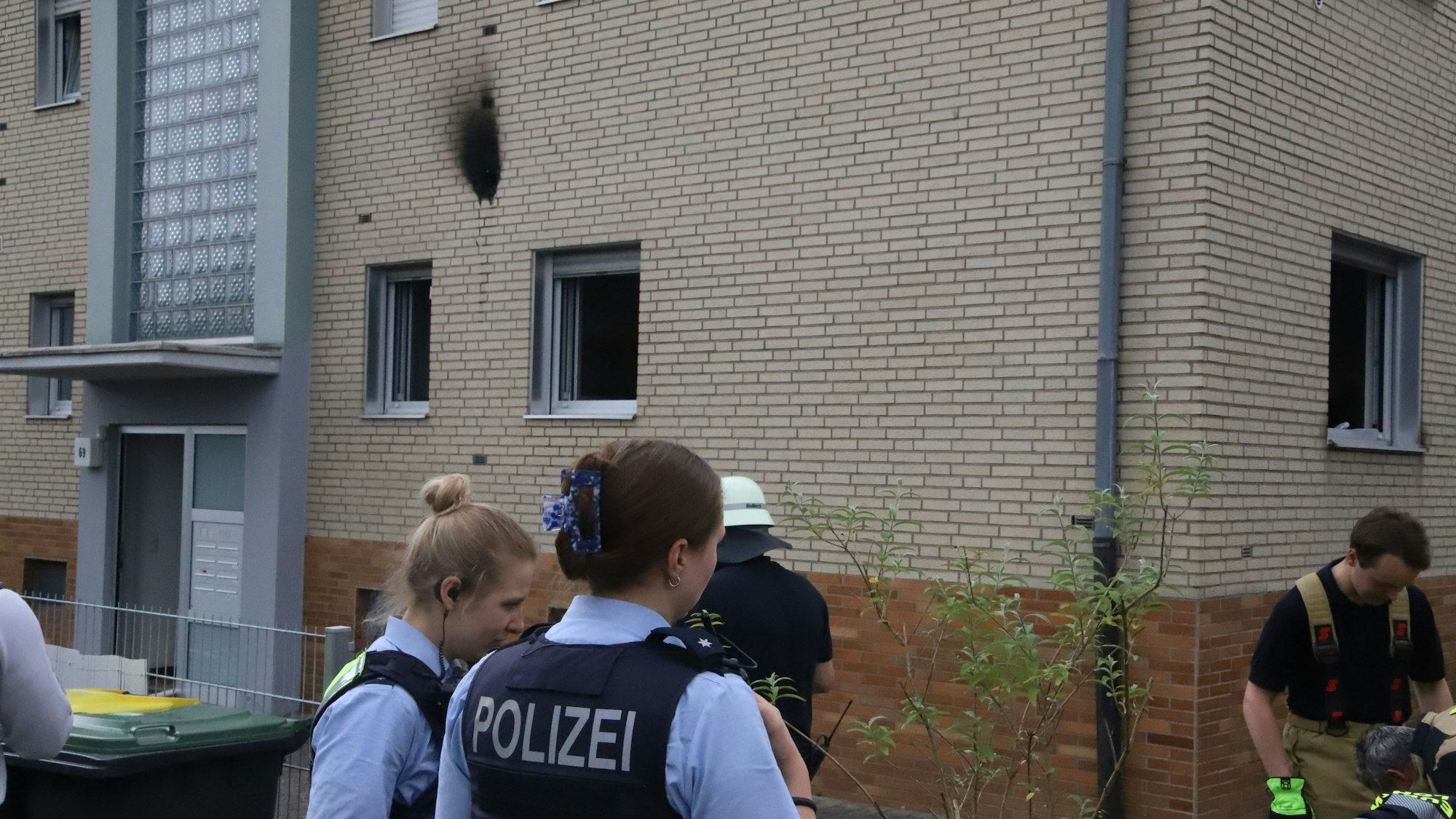 Polizistinnen und Feuerwehrleute stehen vor einem Haus, an dem ein schwarzer Rußfleck zu sehen ist.