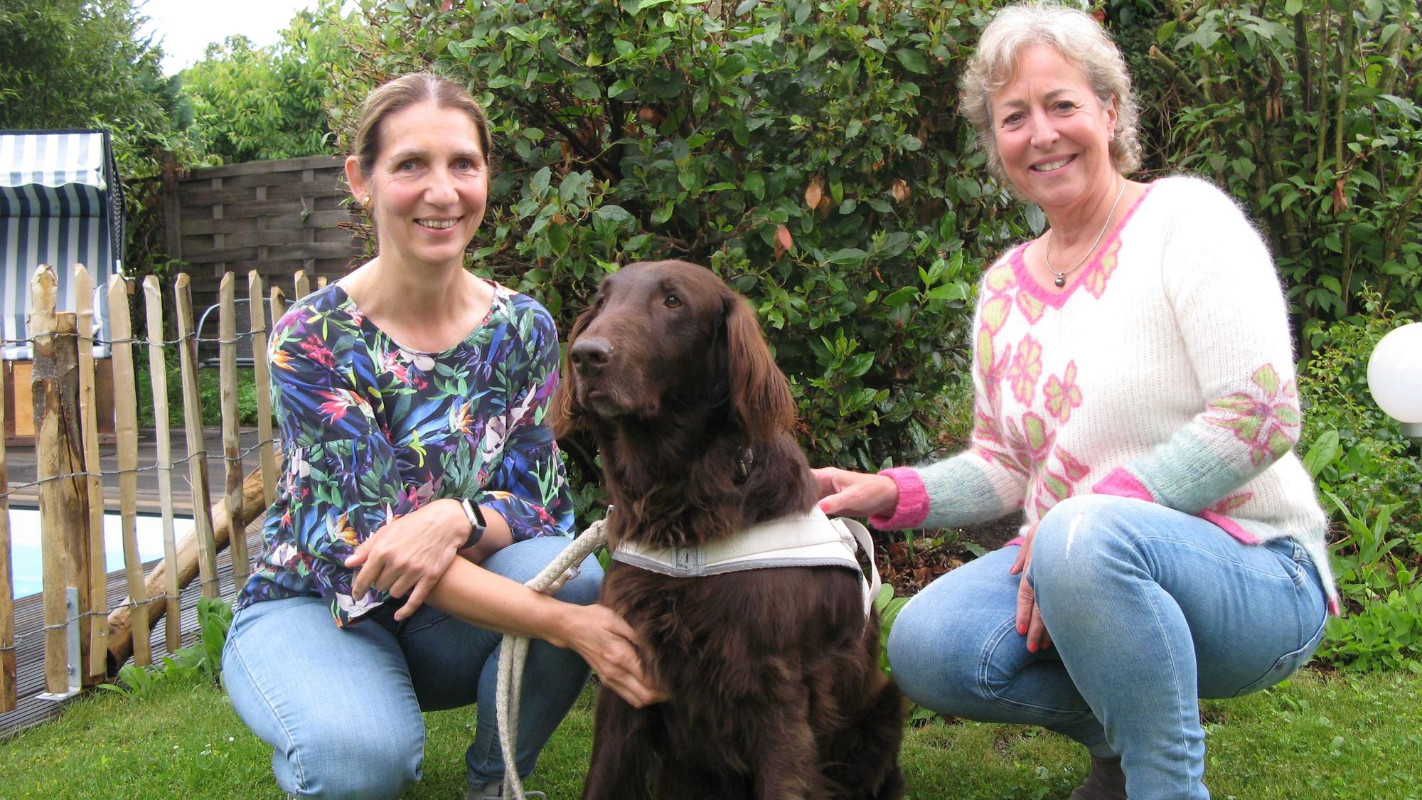 Britta Biersch, Assistenzhund Eido und Barbara Kossack auf einem Rasen im Garten.