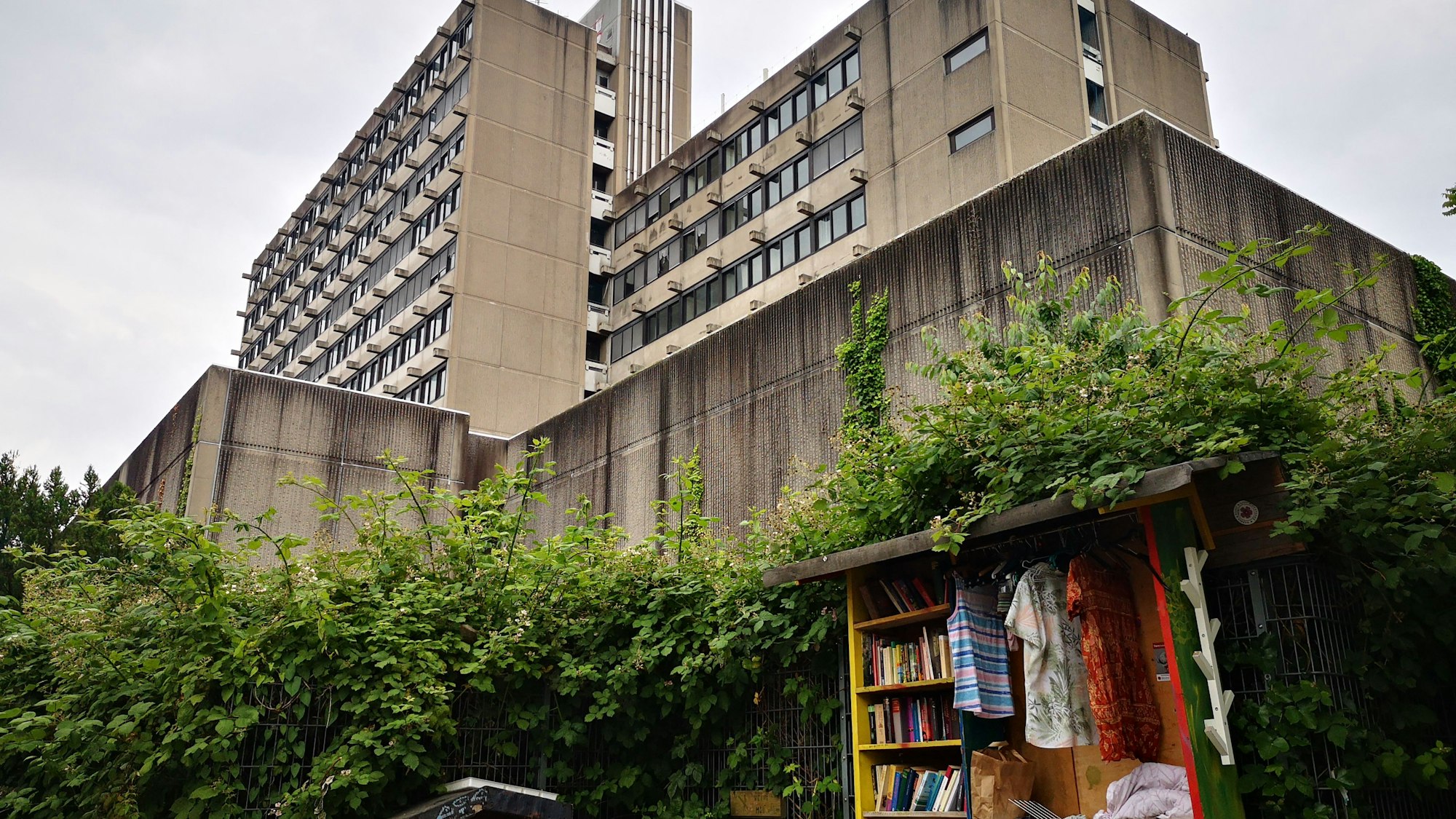 Ein Beton-Hochhaus erhebt sich vor dem grauen Himmel. Direkt davor wachsen Büsche, vor denen ein öffentlicher Tausch-Stand für Passanten aufgestellt ist.