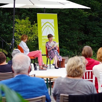 Mariana Leky und Bettina Böttinger sitzen auf einer Bühne, hinter ihnen viel Grün. Vor ihnen schauen die Zuschauer gespannt nach vorne, während Leky aus ihrem Roman „Was man von hier aus sehen kann“ liest.