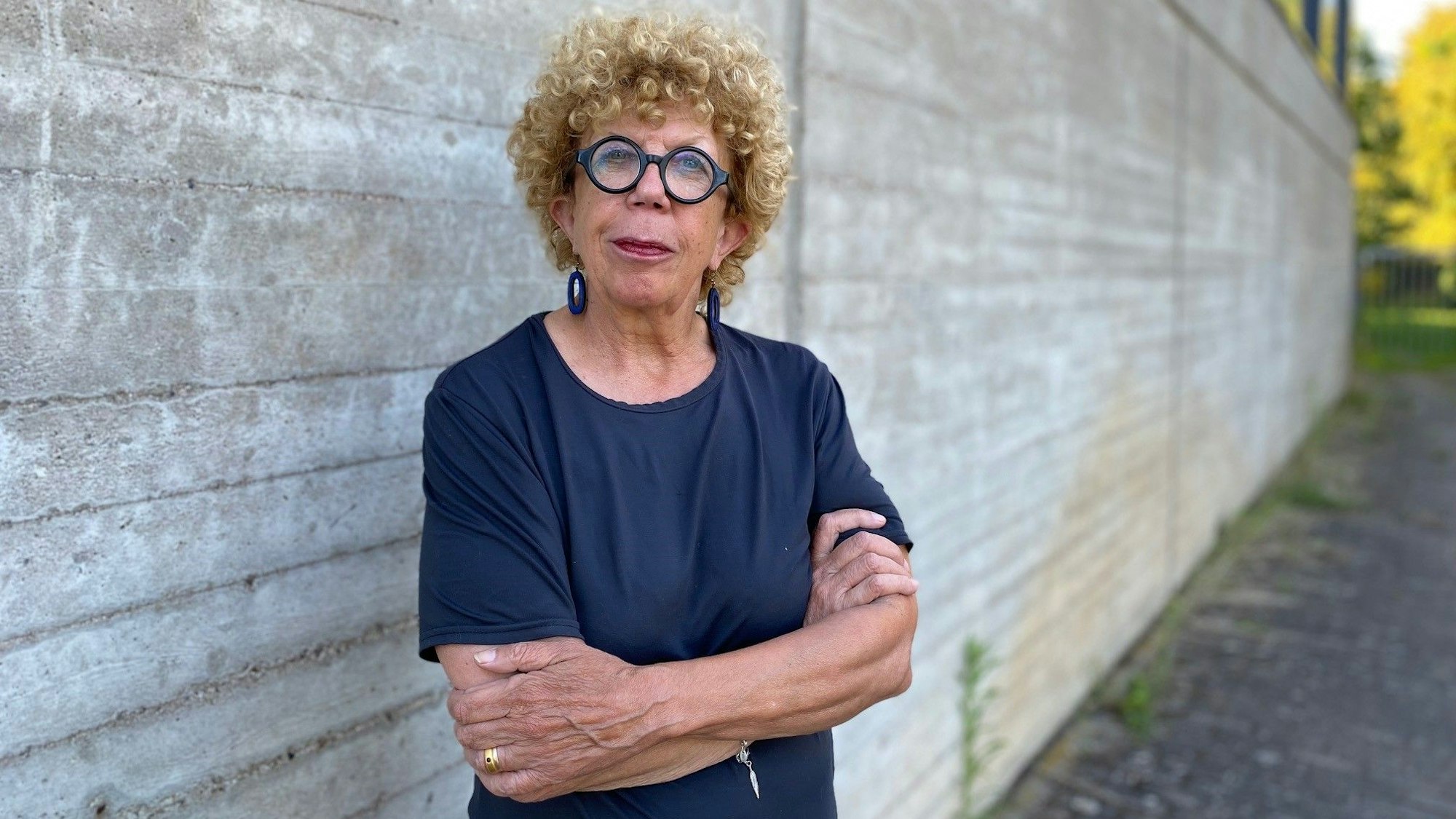 Auf dem Foto ist eine Frau mit lockigem Haar und einer runden Brille zu sehen. Sie steht mit verschränkten Armen vor einer Mauer.