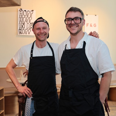Die beiden Köche Grischa Herbig und Caspar Krings sind mit schwarzer Schürze und freundlich lachend in ihrem Restaurant zu sehen.