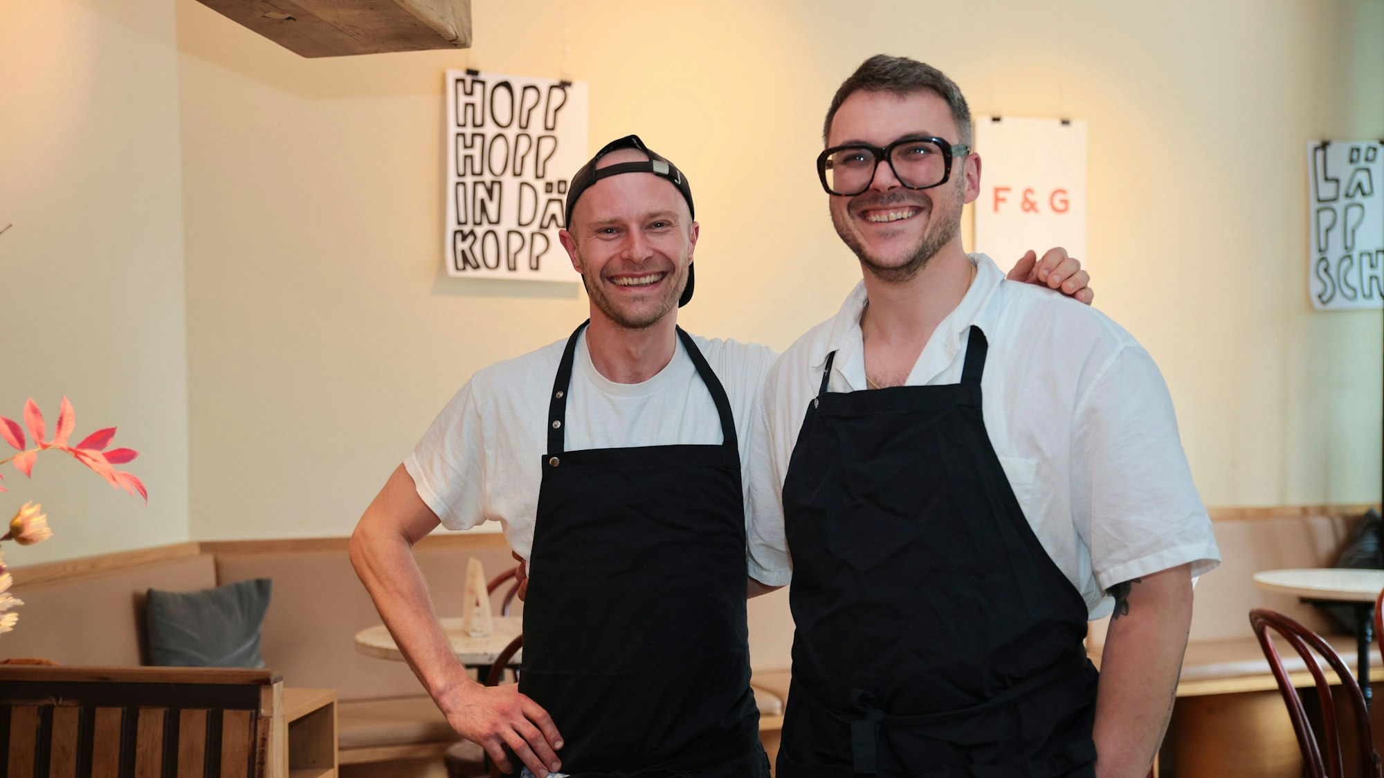 Die beiden Köche Grischa Herbig und Caspar Krings sind mit schwarzer Schürze und freundlich lachend in ihrem Restaurant zu sehen.