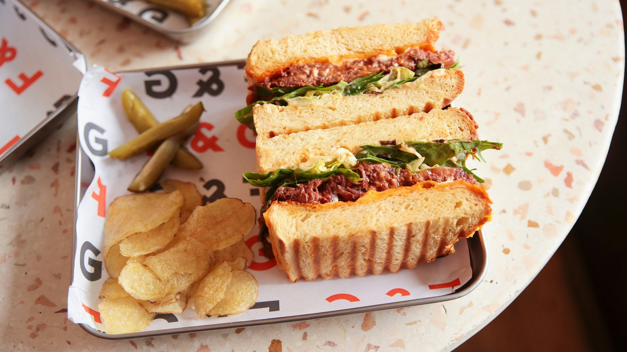Ein Sandwich mit Ossobuco belegt und Kartoffelchips daneben auf einem bunten Schälchen.