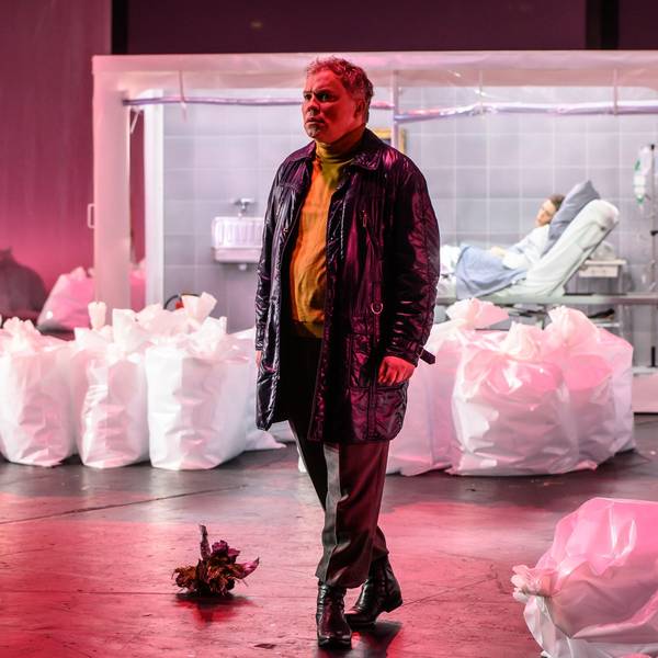 Hagen Matzeit trägt eine Lederjacke und einen gelben Rollkragenpullover. Er ist umgeben von befüllten, aufrecht stehenden Plastiksäcken. Im Hintergrund Kathrin Zukowski in einem Krankenbett, in Quarantäne. Die Bühne ist pink beleuchtet.