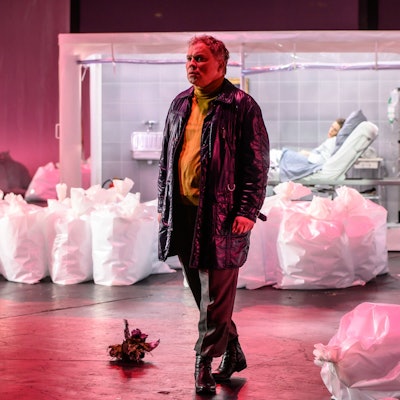 Hagen Matzeit trägt eine Lederjacke und einen gelben Rollkragenpullover. Er ist umgeben von befüllten, aufrecht stehenden Plastiksäcken. Im Hintergrund Kathrin Zukowski in einem Krankenbett, in Quarantäne. Die Bühne ist pink beleuchtet.