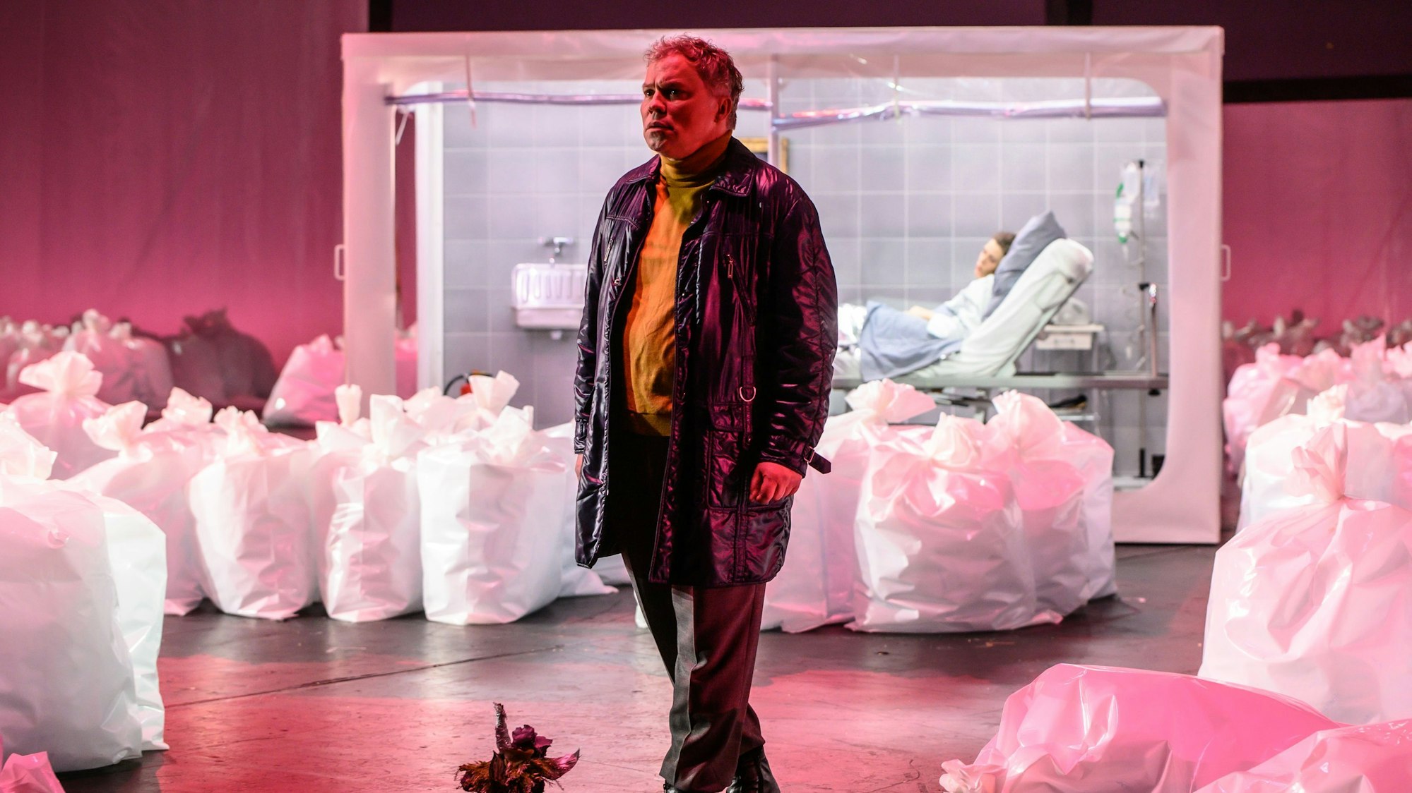 Hagen Matzeit trägt eine Lederjacke und einen gelben Rollkragenpullover. Er ist umgeben von befüllten, aufrecht stehenden Plastiksäcken. Im Hintergrund Kathrin Zukowski in einem Krankenbett, in Quarantäne. Die Bühne ist pink beleuchtet.