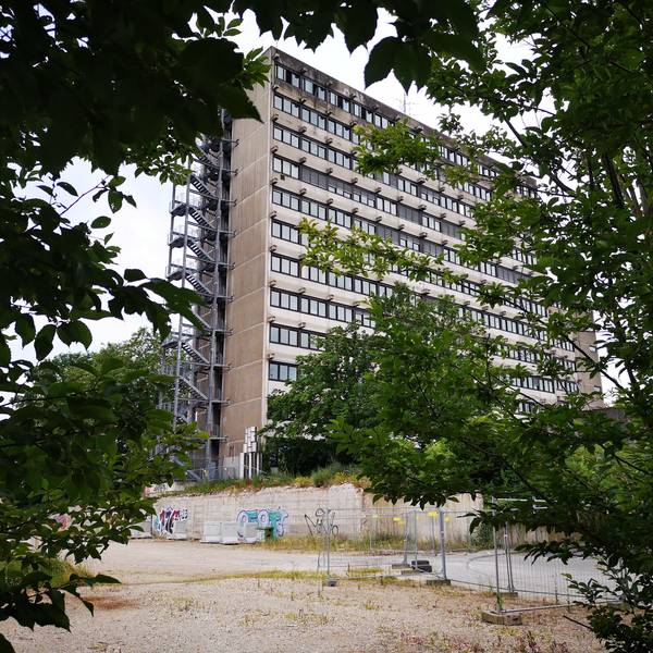 Ein graues Hochhaus mit vielen Fenstern und einer Gittertreppe an der linken Seite ist von Bäumen und einer Brachfläche umgeben.