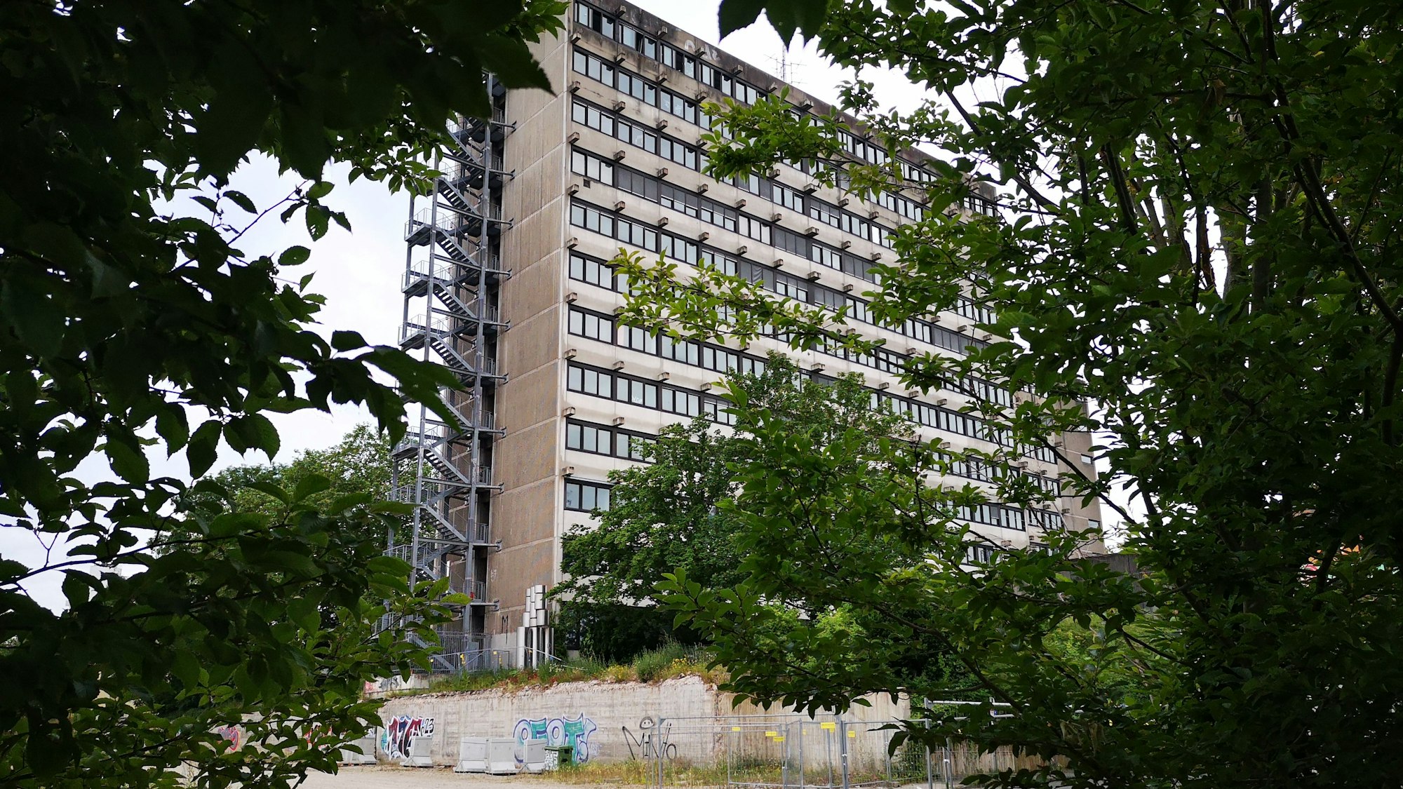 Ein graues Hochhaus mit vielen Fenstern und einer Gittertreppe an der linken Seite ist von Bäumen und einer Brachfläche umgeben.