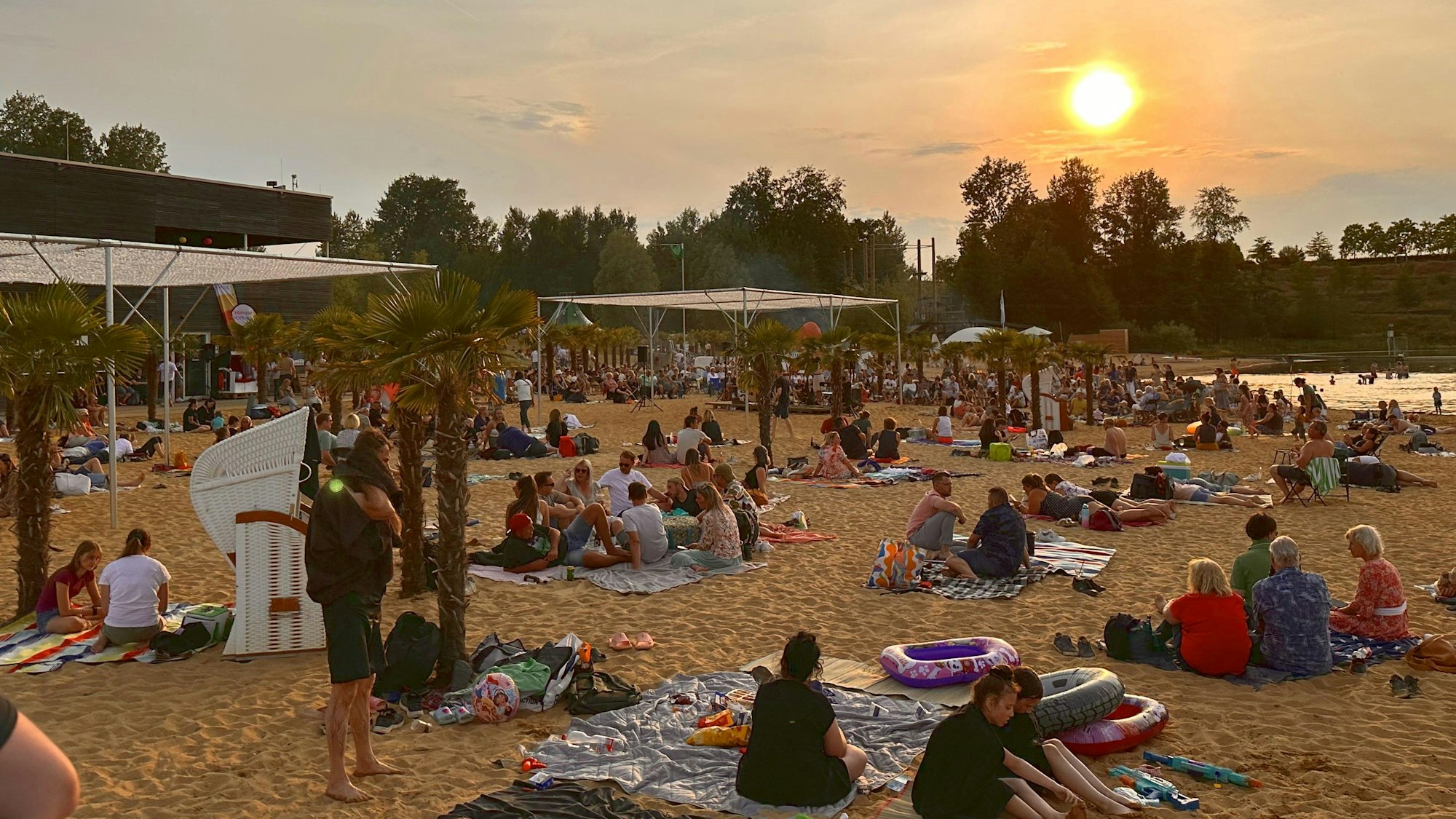 Viele Menschen sitzen beim Sonnenuntergang am Sandstrand des Zülpicher Sees.