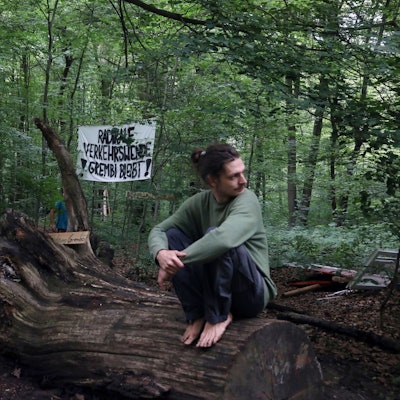 Ein Mann hockt barfuß auf einem Baumstumpf im Gremberger Wäldchen, im Hintergrund ein Protestplakat gegen den Ausbau der Autobahn 4.