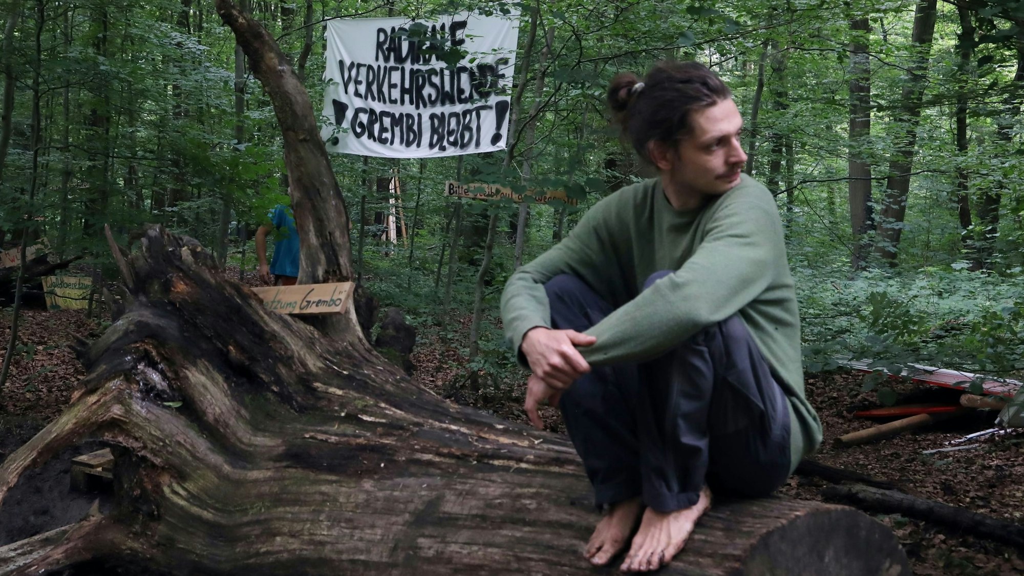 Ein Mann hockt barfuß auf einem Baumstumpf im Gremberger Wäldchen, im Hintergrund ein Protestplakat gegen den Ausbau der Autobahn 4.