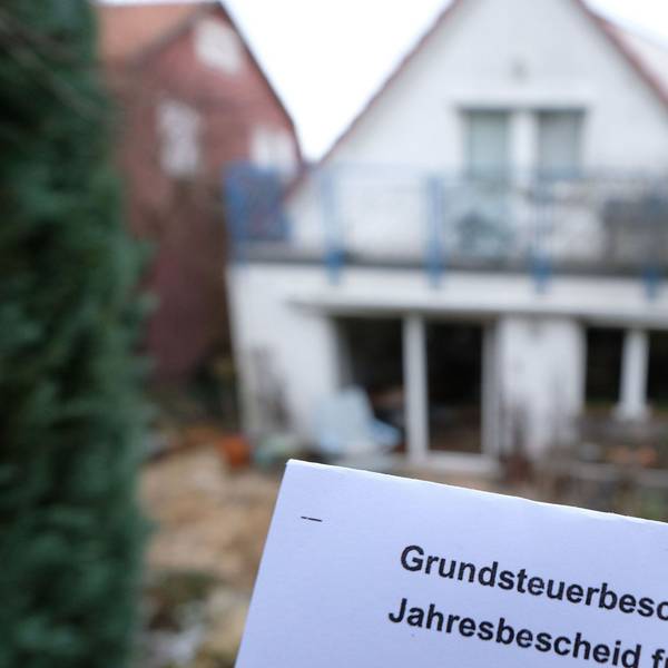 Ein Grundsteuerbescheid für 2024 wird vor einem Privatgrundstück hochgehalten.