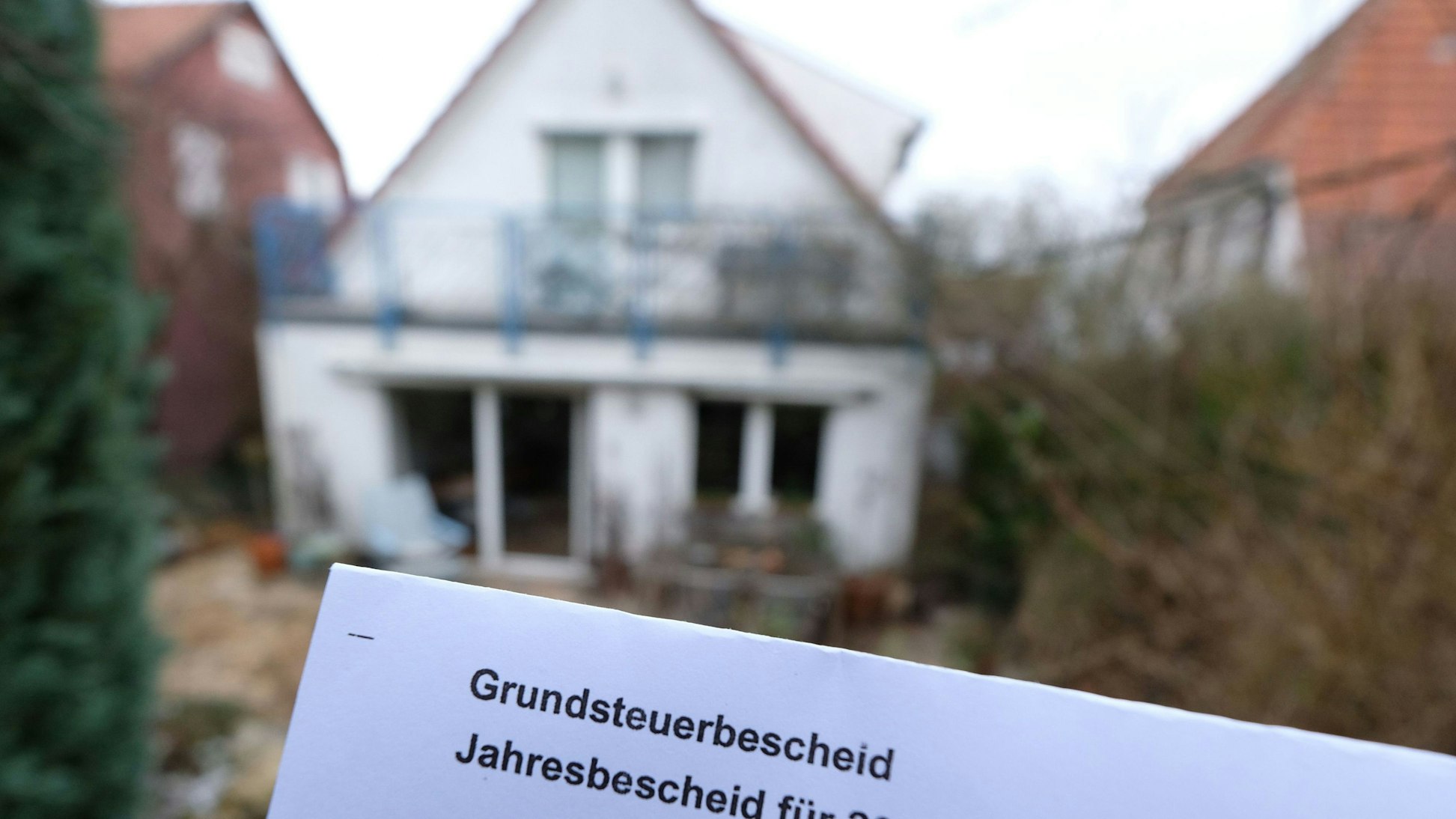 Ein Grundsteuerbescheid für 2024 wird vor einem Privatgrundstück hochgehalten.