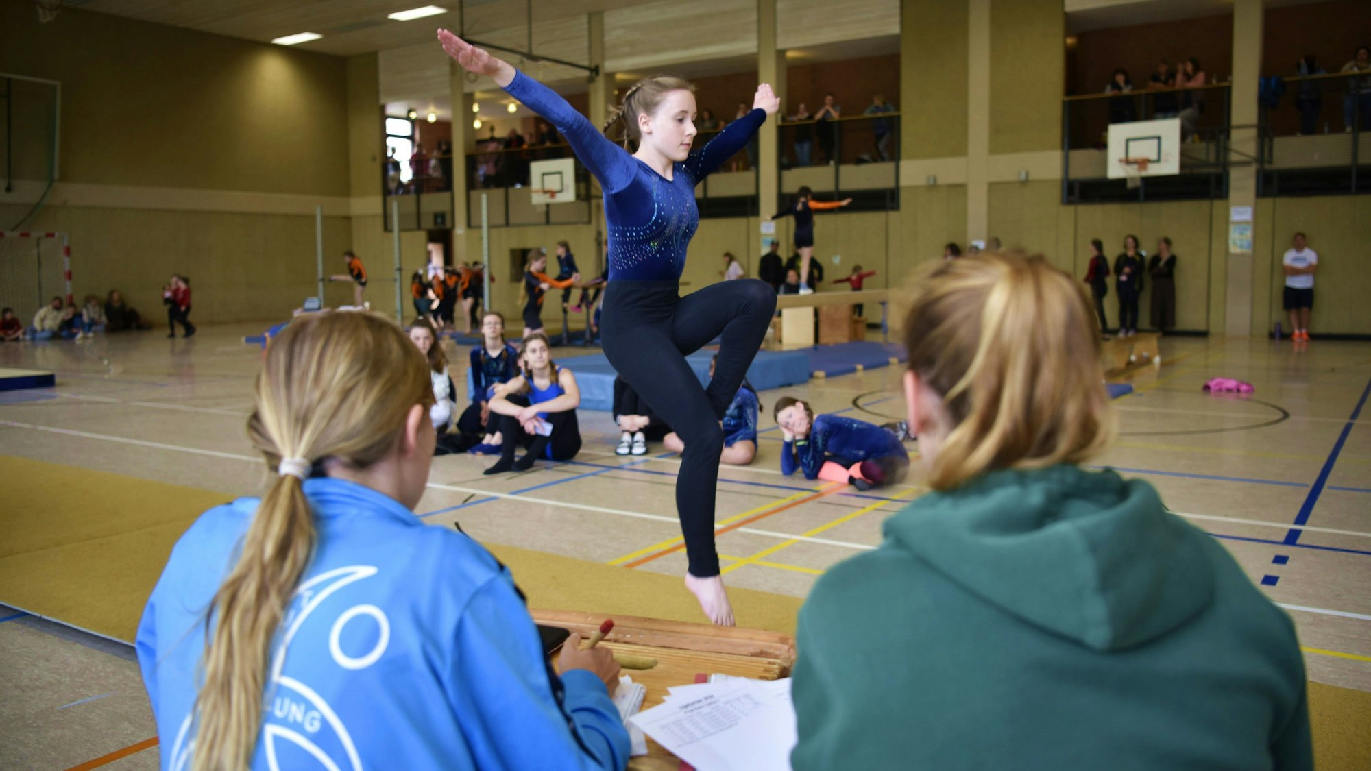 Eine Turnerin des Tus Schleiden zeigt beim Liga-Turnen ihre Bodenübung. Sie macht gerade einen Pferdchensprung und wird dabei von zwei Kampfrichterinnen sowie anderen Turnerinnen beobachtet.