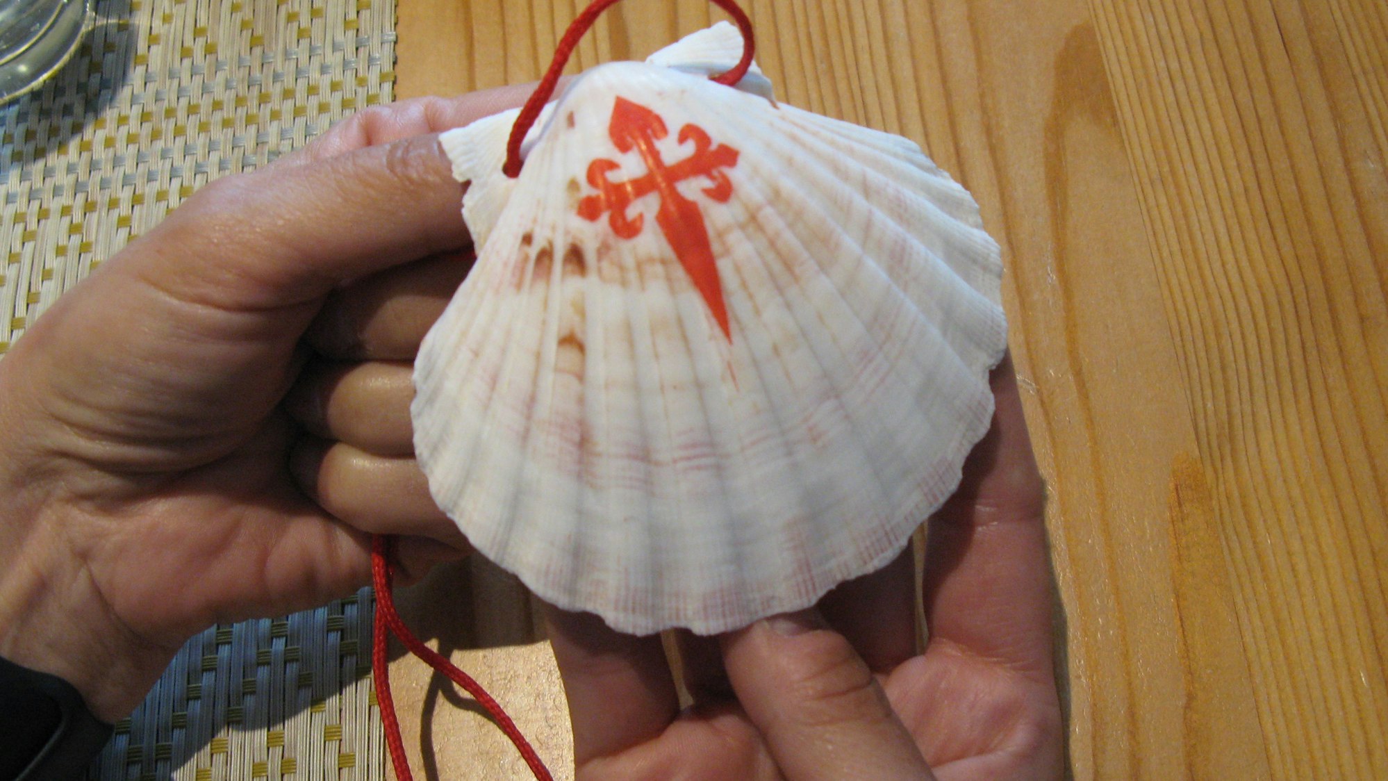 Eine Jakobsmuschel mit rotem Symbol, gehalten von zwei Händen.