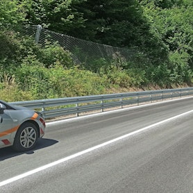 Eine leicht geschwungene Straße mit neuer Markierung und neuen Leitplanken, links ist ein dicht bewachsener Steilhang mit Steinfangzaun.