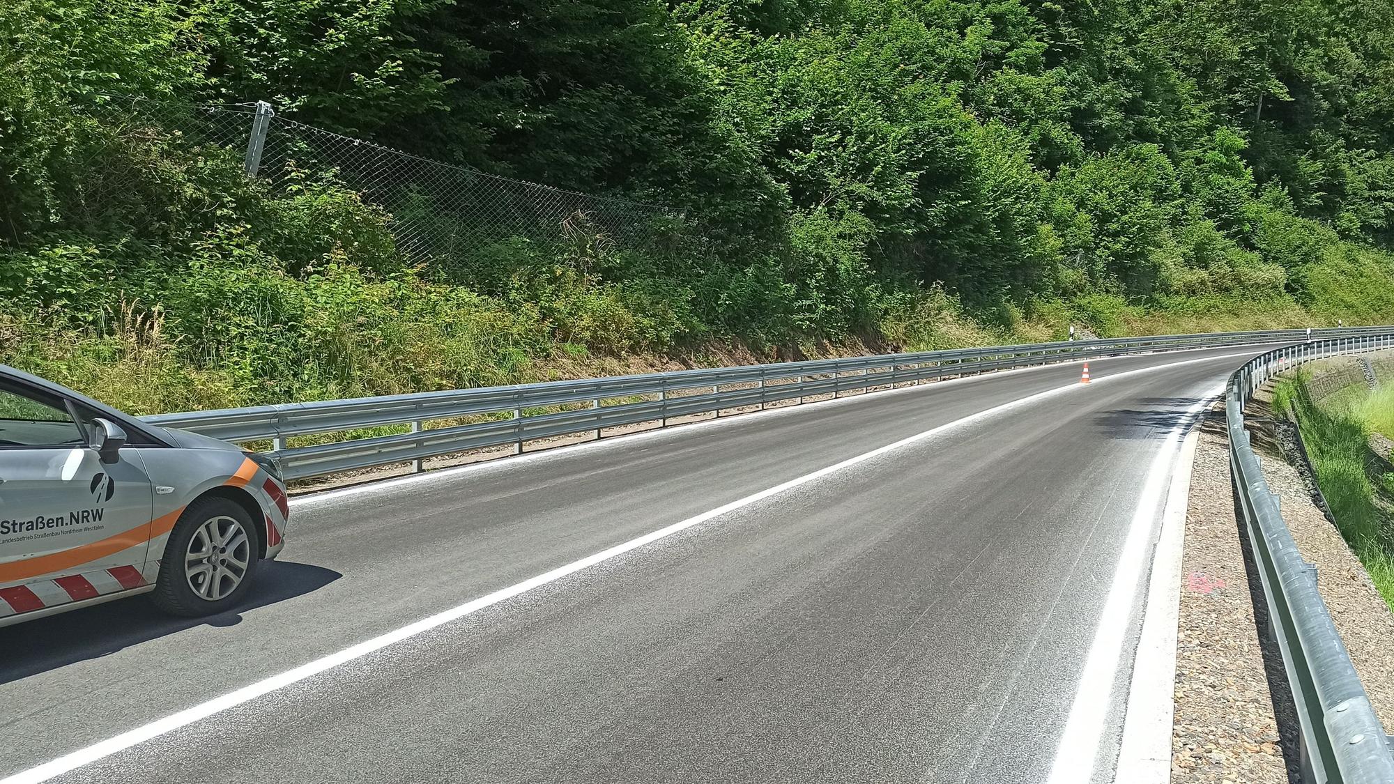 Eine leicht geschwungene Straße mit neuer Markierung und neuen Leitplanken, links ist ein dicht bewachsener Steilhang mit Steinfangzaun.