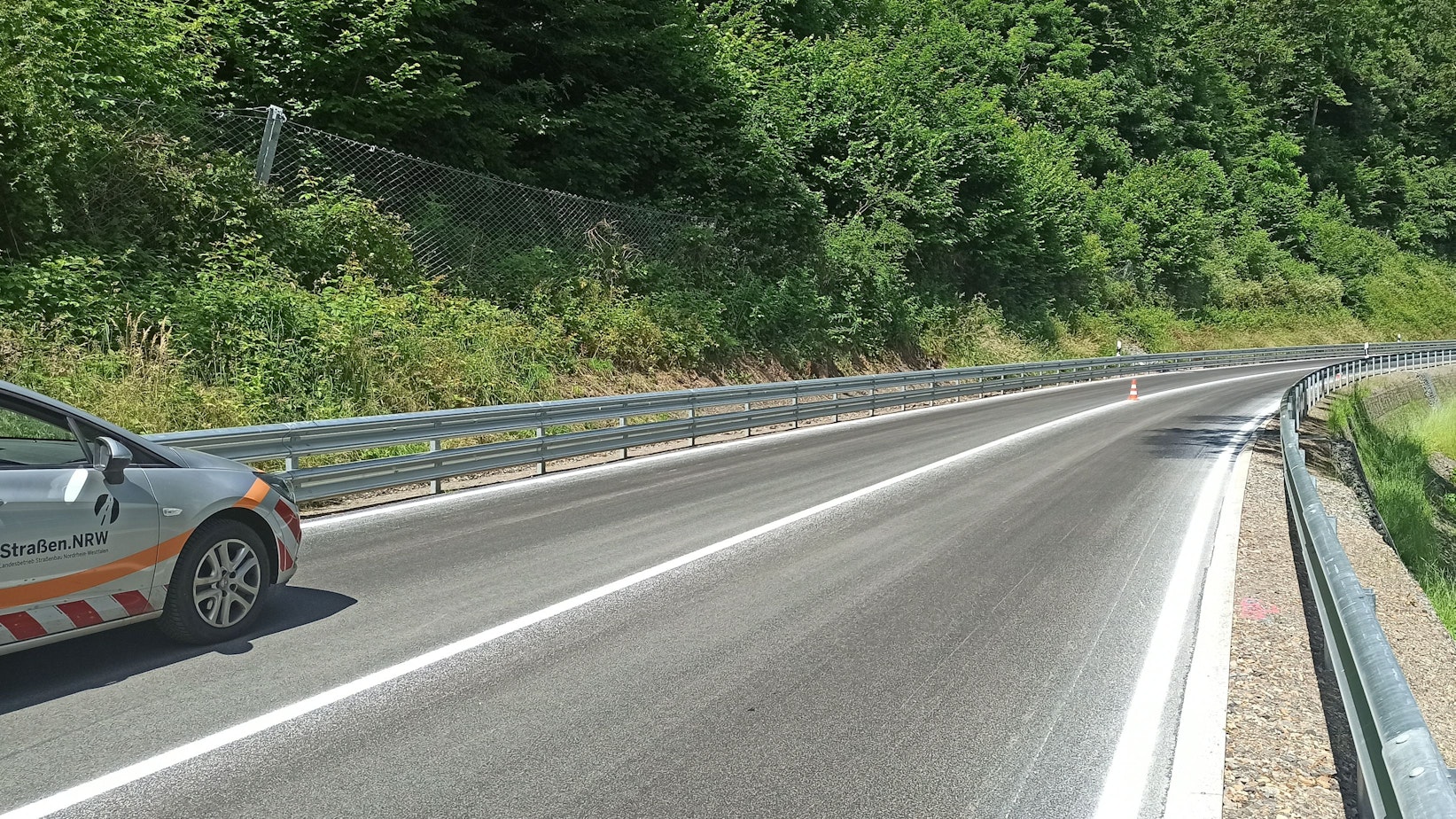 Eine leicht geschwungene Straße mit neuer Markierung und neuen Leitplanken, links ist ein dicht bewachsener Steilhang mit Steinfangzaun.