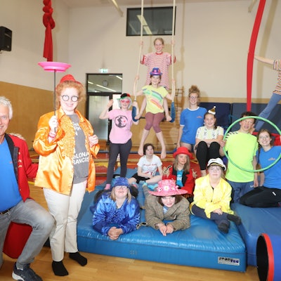 12 junge, verkleidete Artistinnen und Artisten des inklusiven „Sternschnuppen“-Kurses sitzen, liegen und stehen auf einer blauen Trainingsmatte, drei hängen in einem Seil darüber