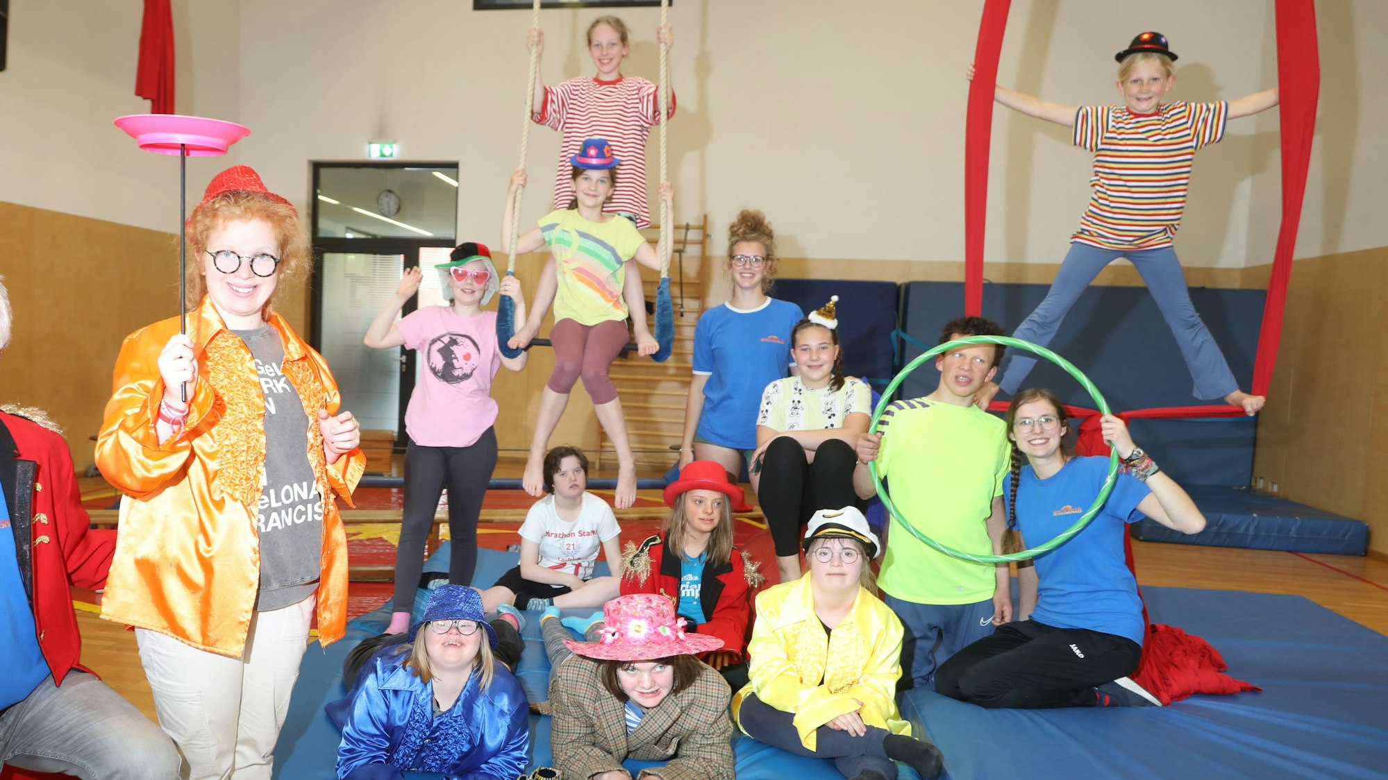 12 junge, verkleidete Artistinnen und Artisten des inklusiven „Sternschnuppen“-Kurses sitzen, liegen und stehen auf einer blauen Trainingsmatte, drei hängen in einem Seil darüber
