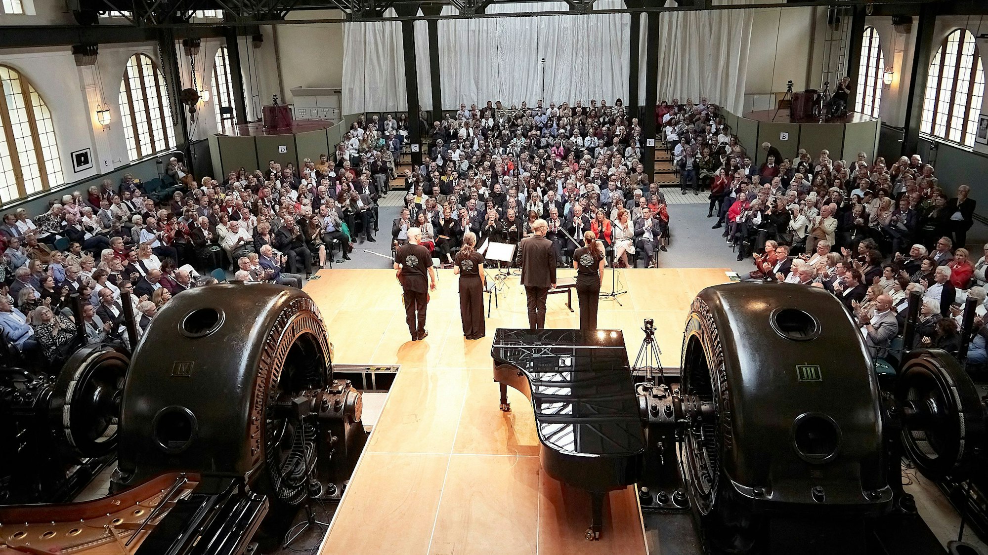Das Bild zeigt den Konzertsaal im Jugendstilkraftwerk aus der Vogelperspektive. Zusehen sind Turbinen, vier Musiker, die davor auf einer Bühne stehen und ein großes Publikum.