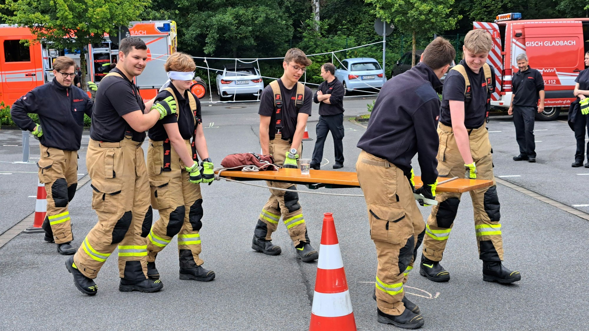 Feuerwehrleute tragen einen Tisch mit untergelegten Äxten. Eine Feuerwehrfrau hat die Augen verbunden und wird geführt.