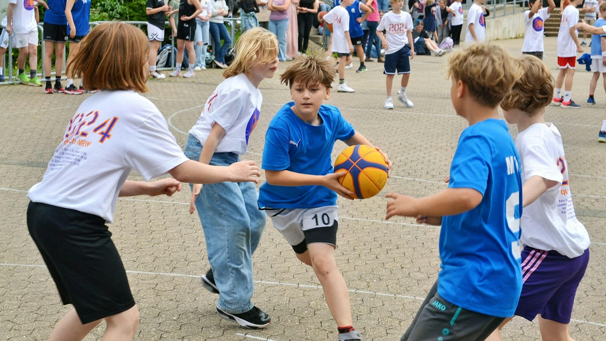 Mehrere Kinder spielen zusammen Basketball. Ein Junge mit blauem t-Shirt hat den Ball in den Händen.