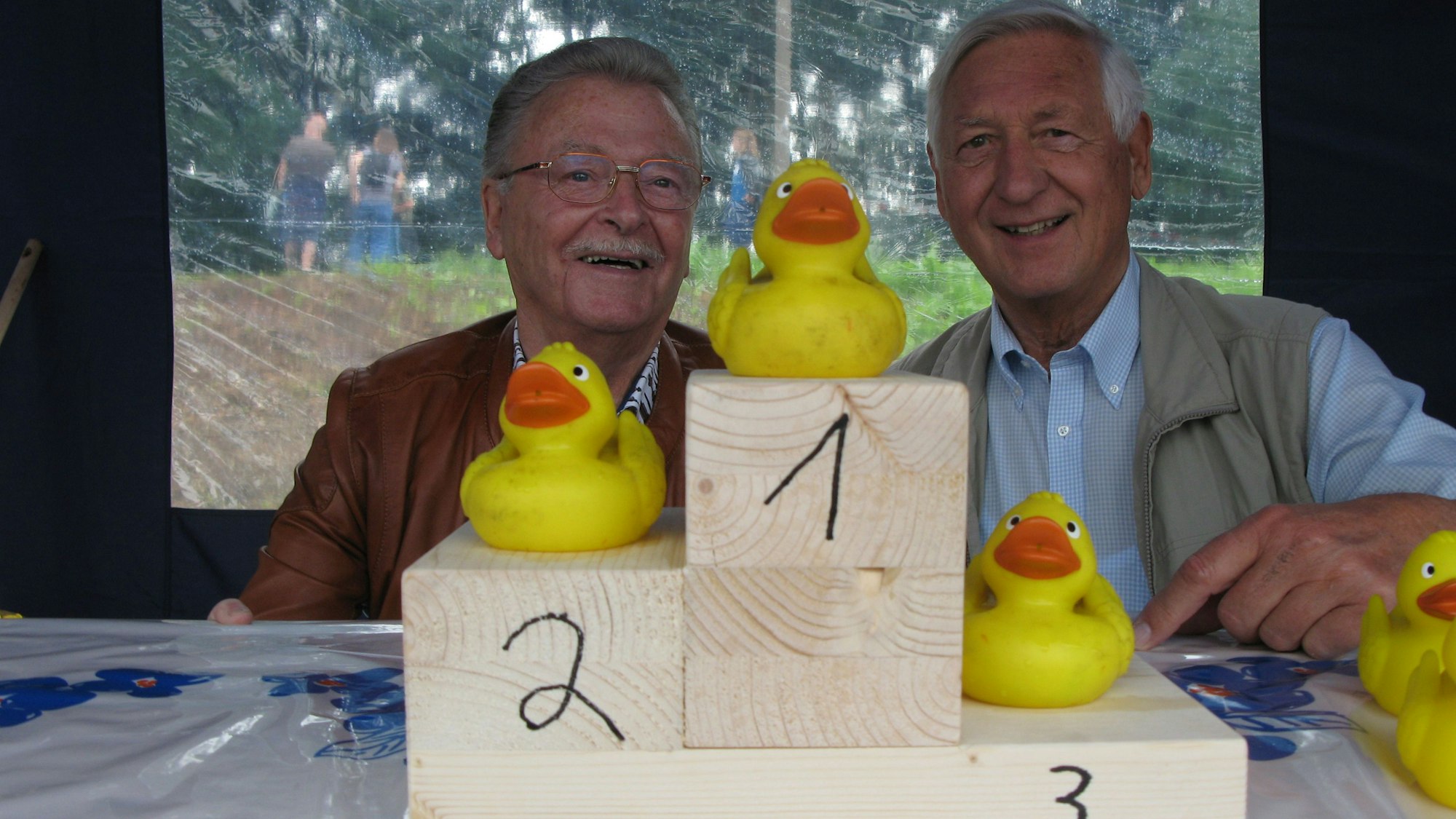 Zwei Männer hinter dem Siegertreppchen aus Holz, auf dem drei gelbe Plastikenten platziert sind.