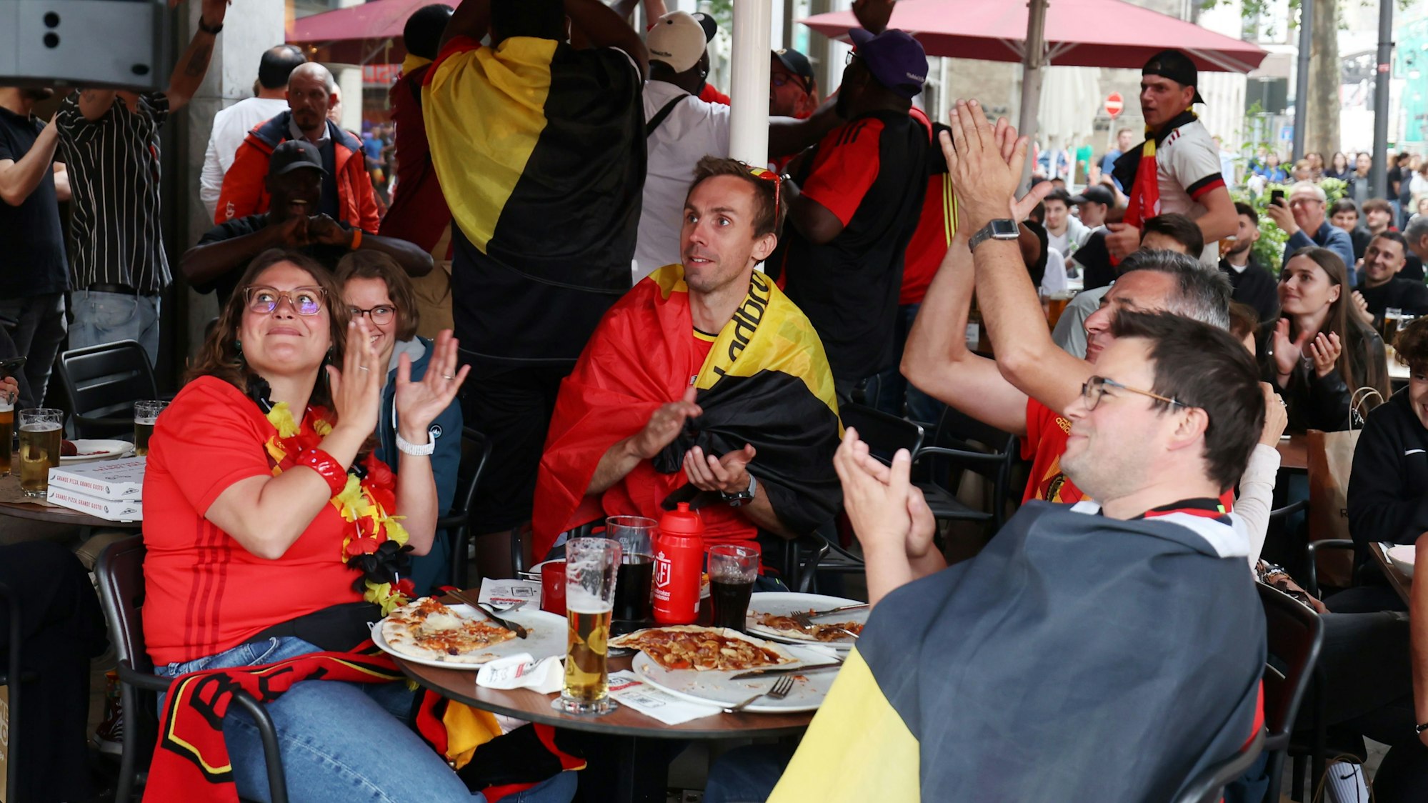 Belgische Fans sitzn an einem Tisch zusammen und schauen das Spiel auf der Leinwand.