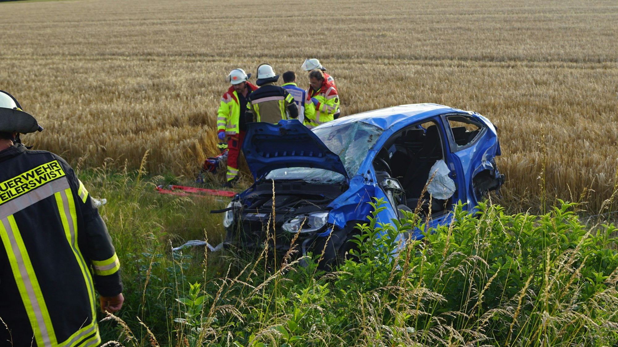 Rettungskräfte stehen an einem zerstörten blauen Auto an einem Feld.