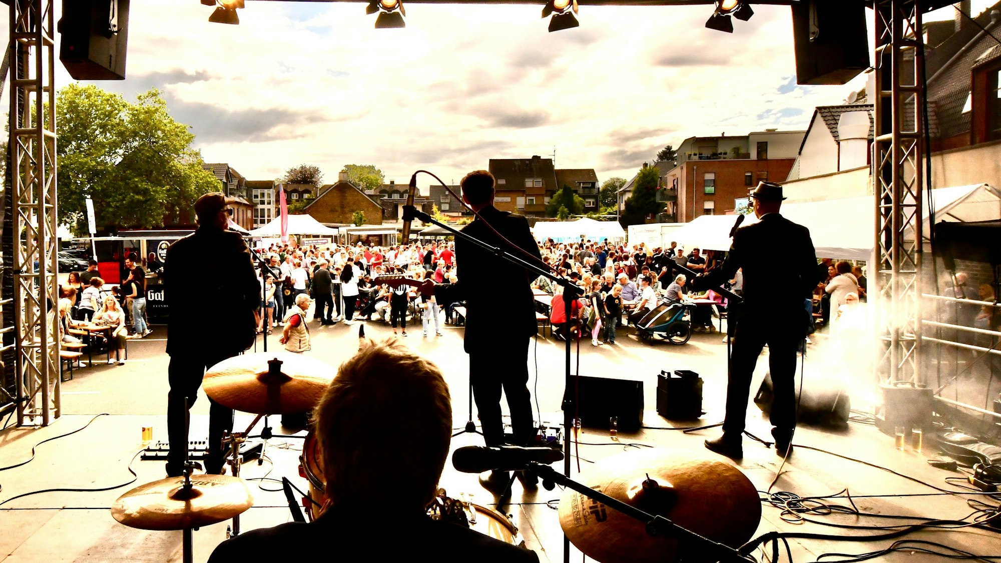 Im Vordergrund vier Musiker auf einer Bühne, im Hintergrund sind ein viele Menschen zu sehen.