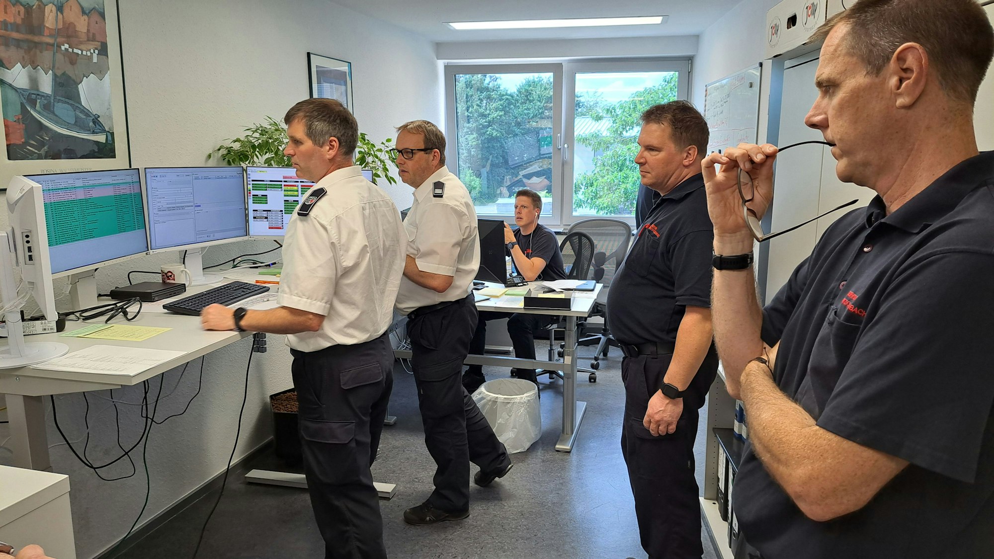 Zwei Leitstellenmitarbeiter in weißen Hemden stehen vor Bildschirmen und disponieren, weitere Feuerwehrleute schauen zu.