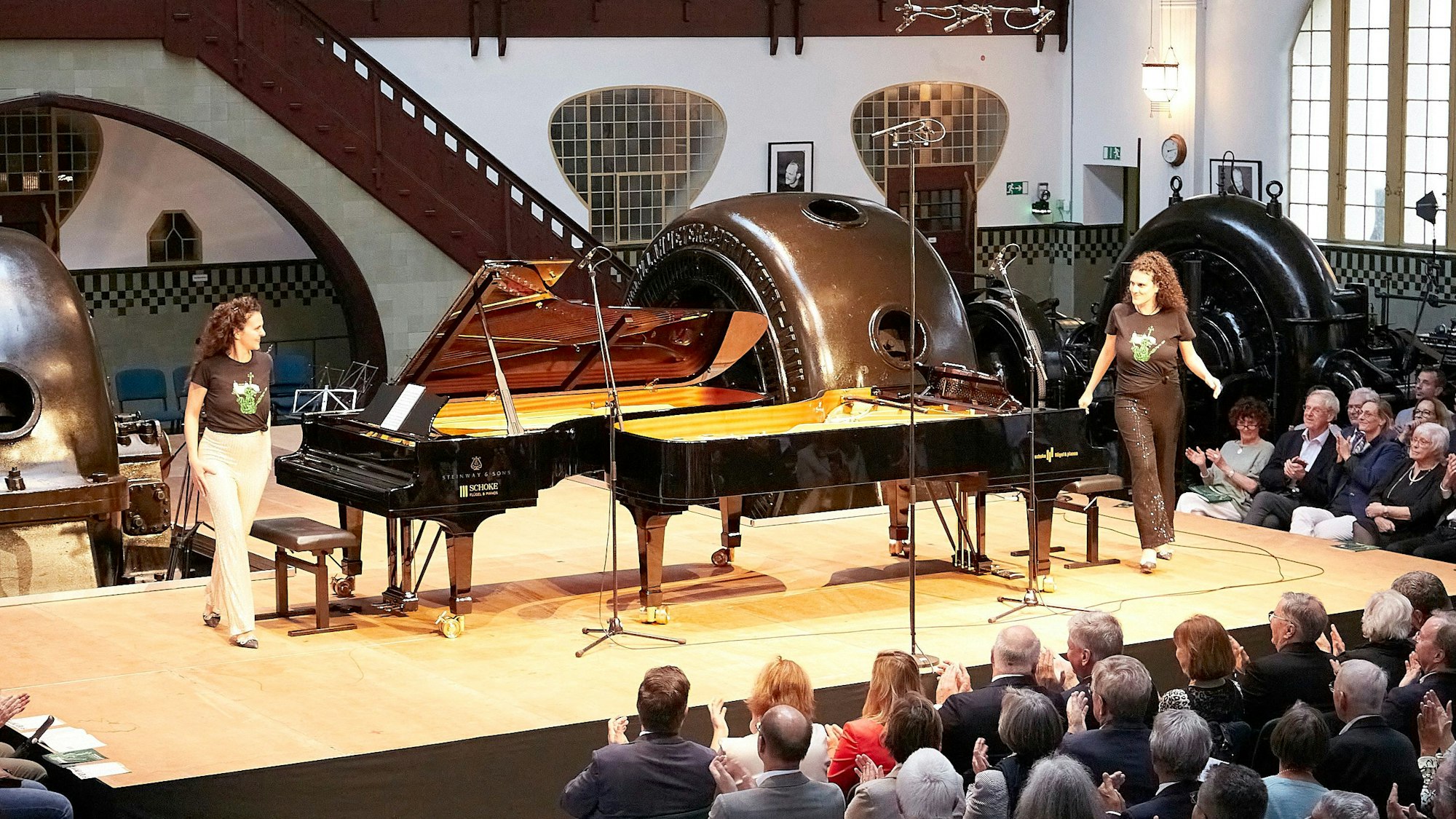 Die Schwestern Danae und Kiveli Dörken stehen neben zwei Flügeln auf einer Bühne, im Hintergrund sind Turbinen erkennbar.