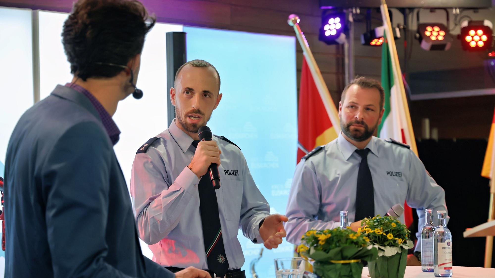 Zwei Polizisten in Uniform stehen an einem Stehtisch und sprechen mit einem Moderator.