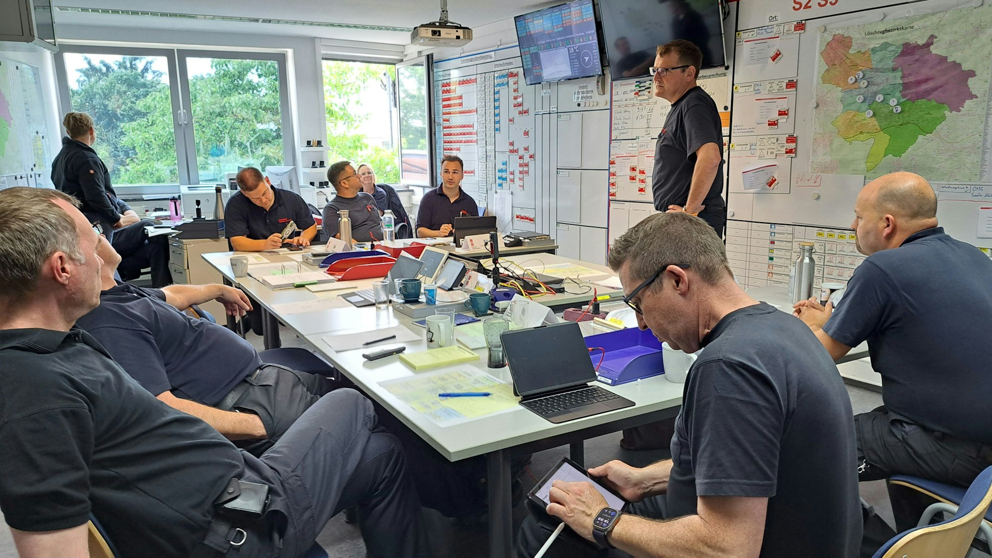 Feuerwehrleute sitzen um einen Tisch, an der Wand sind Bildschirme, Tafeln und Karten zu sehen.