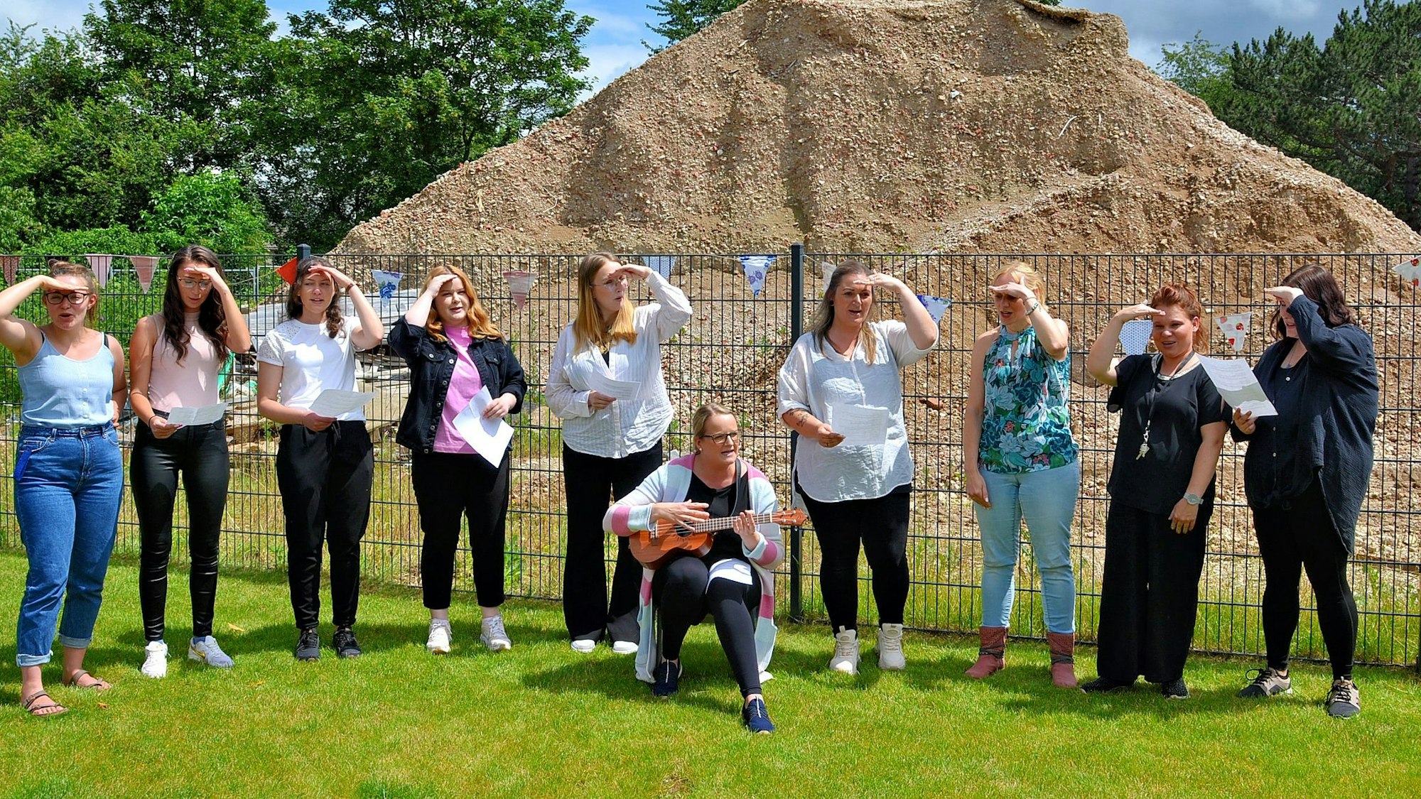 Neun Frauen stehen auf einer Wiese und singen ein Lied. Eine Hand halten sie sich an die Stirn. Eine weitere Frau sitzt auf einem Stuhl und spielt Gitarre.