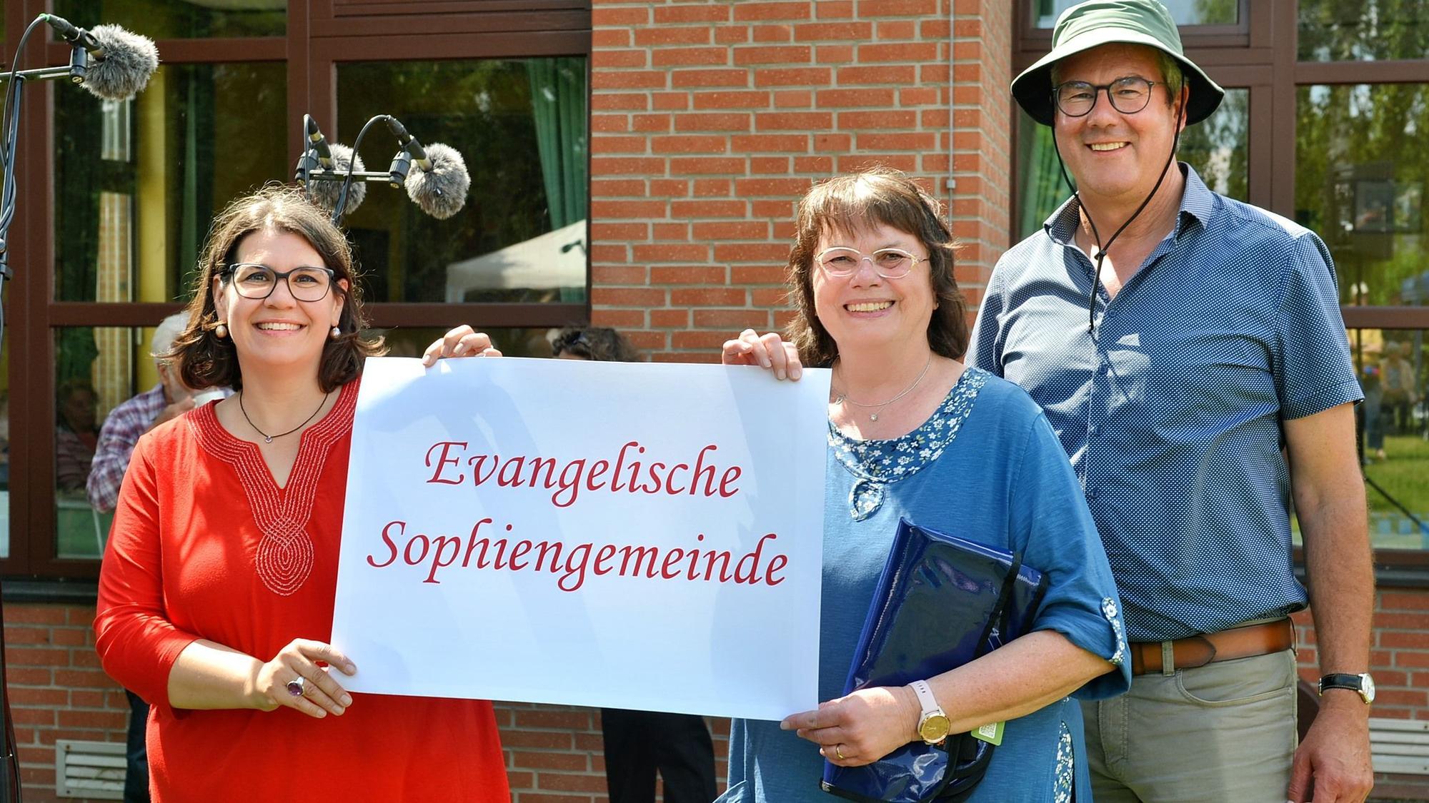 Zwei Frauen halten ein Schild mit der Aufschrift „Evangelische Sophiengemeinde“ fest, daneben steht ein Mann, der einen grünen Anglerhut trägt.