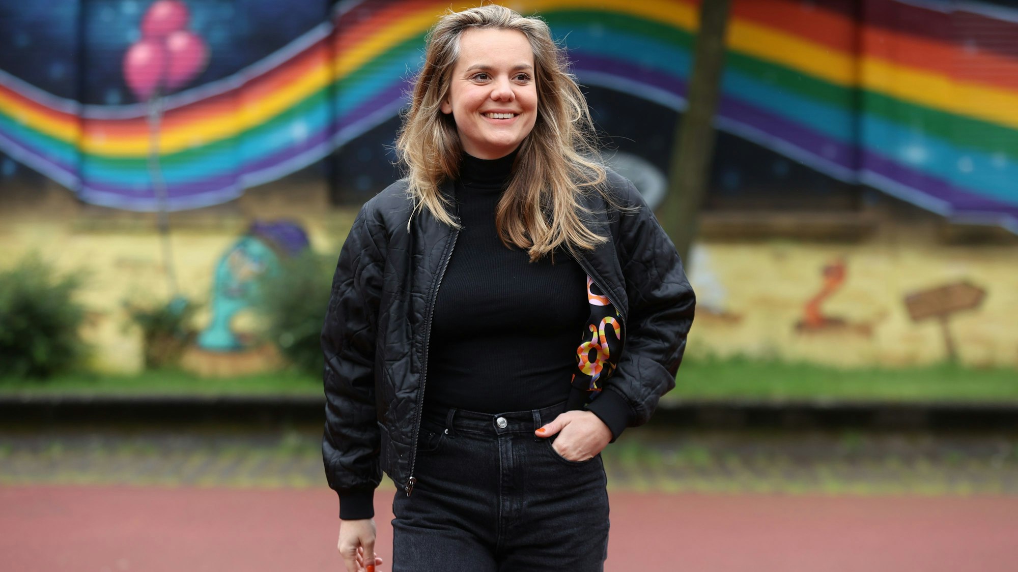 Ricarda Hofman steht vor einer Mauer auf die ein Regenbogen gemalt ist.