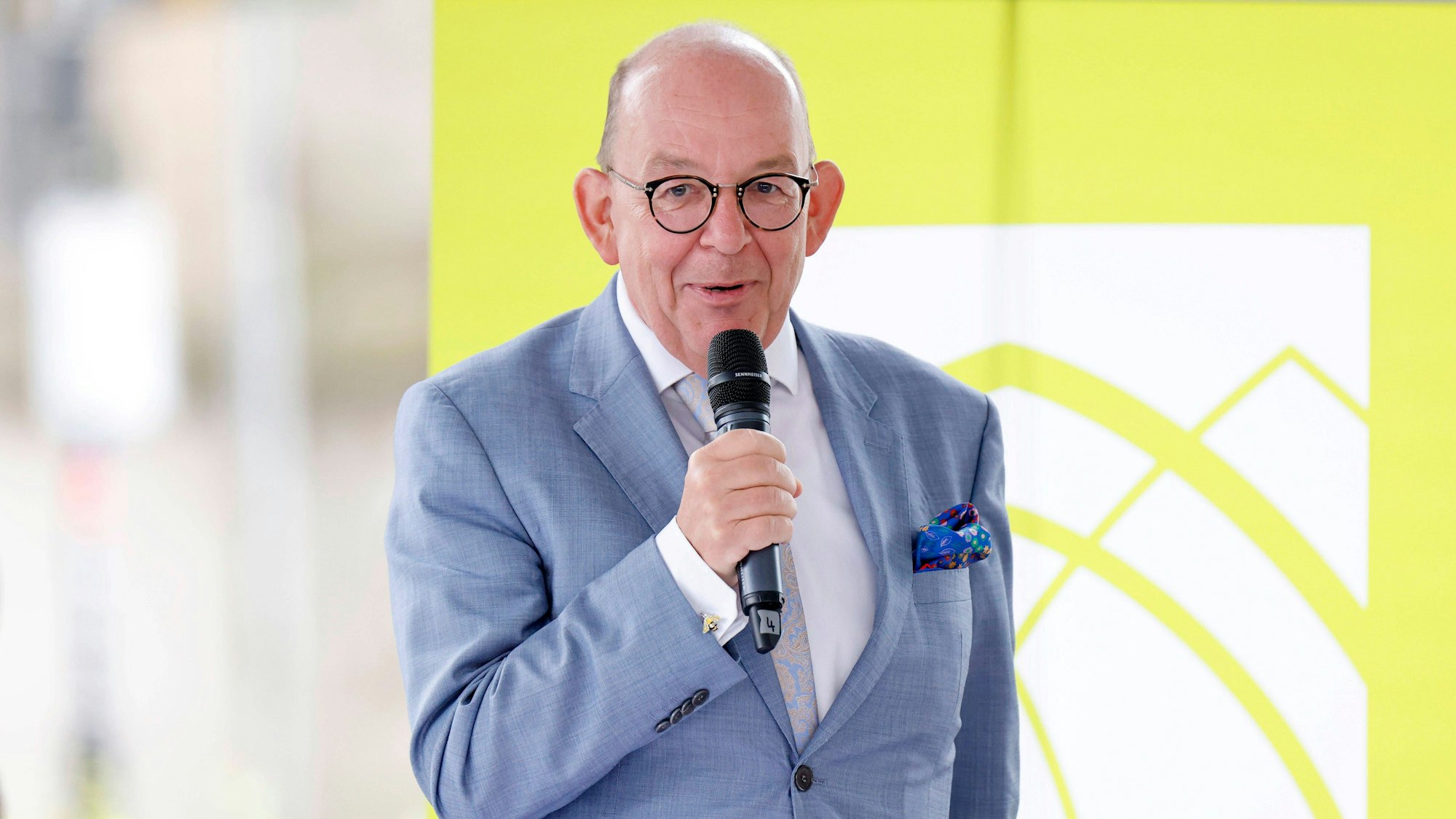 Denis Scheck trägt einen graublauen Anzug und hält ein Mikrofon in der Hand. Er trägt eine runde Brille und hat ein buntes Taschentuch im Revers. Im Hintergrund ein gelbgrünes Banner mit weißem Logo.
