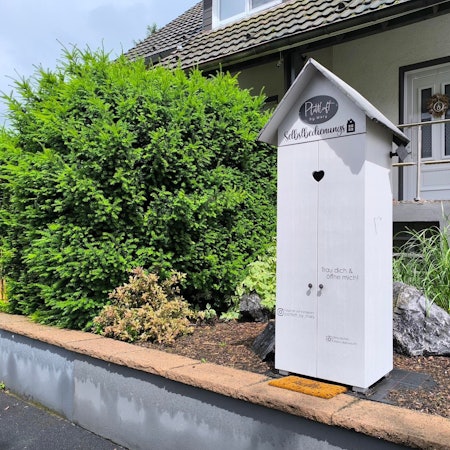 Das Bild zeigt einen Selbstbedienungsladen vor einem Zweifamilienhaus. Um das Häuschen herum stehen viele Büsche.