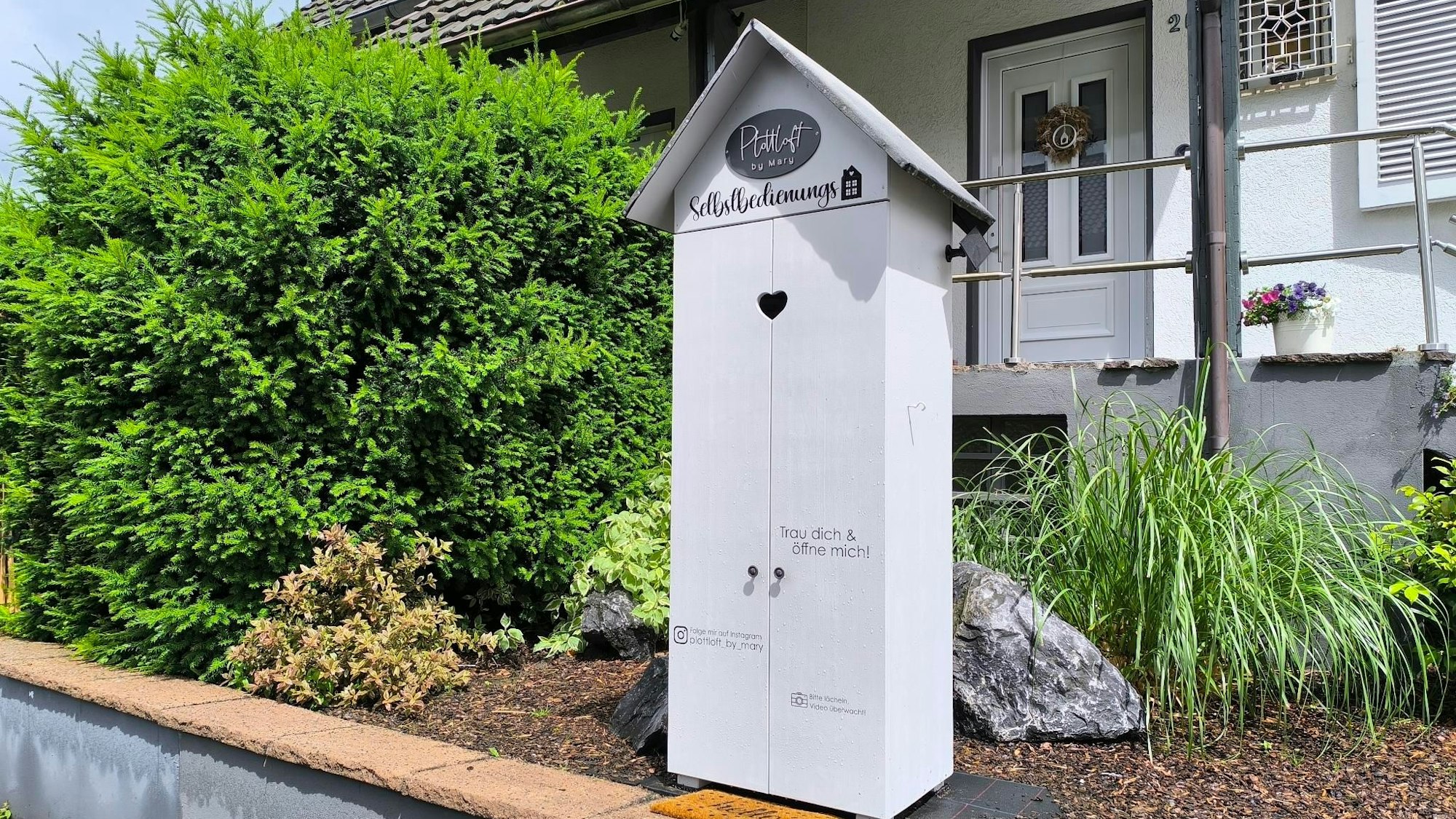 Das Bild zeigt einen Selbstbedienungsladen vor einem Zweifamilienhaus. Um das Häuschen herum stehen viele Büsche.