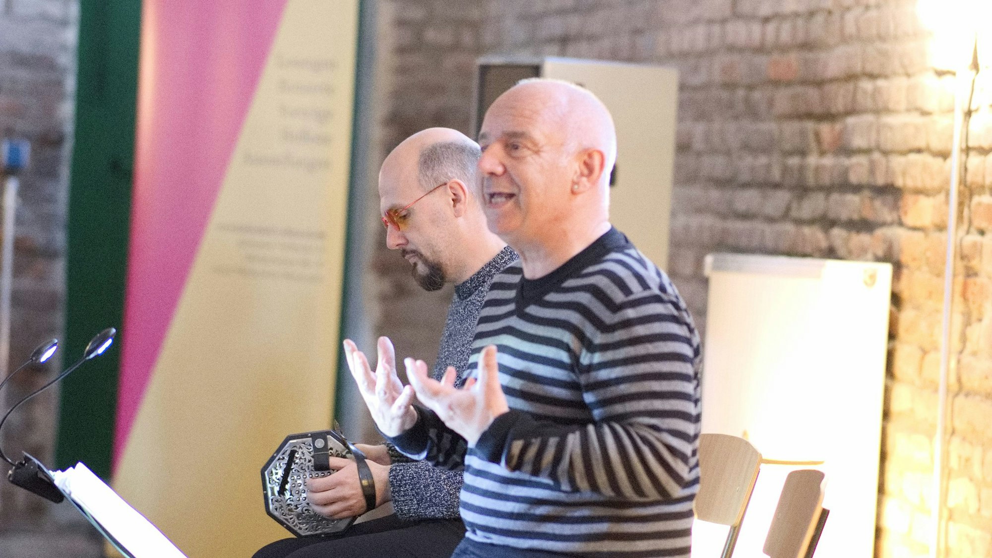 Michael Klevenhaus sitzt gestikulierend auf einem Stuhl, neben ihm musiziert Thomas Zöller auf einer Concertina.