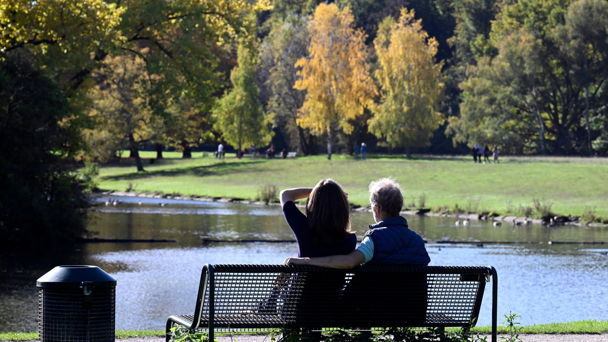 Spaziergänger nutzen den Sonnenschein für eine Pause auf einer Parkbank im Kölner Grüngürtel am Decksteiner Weiher.