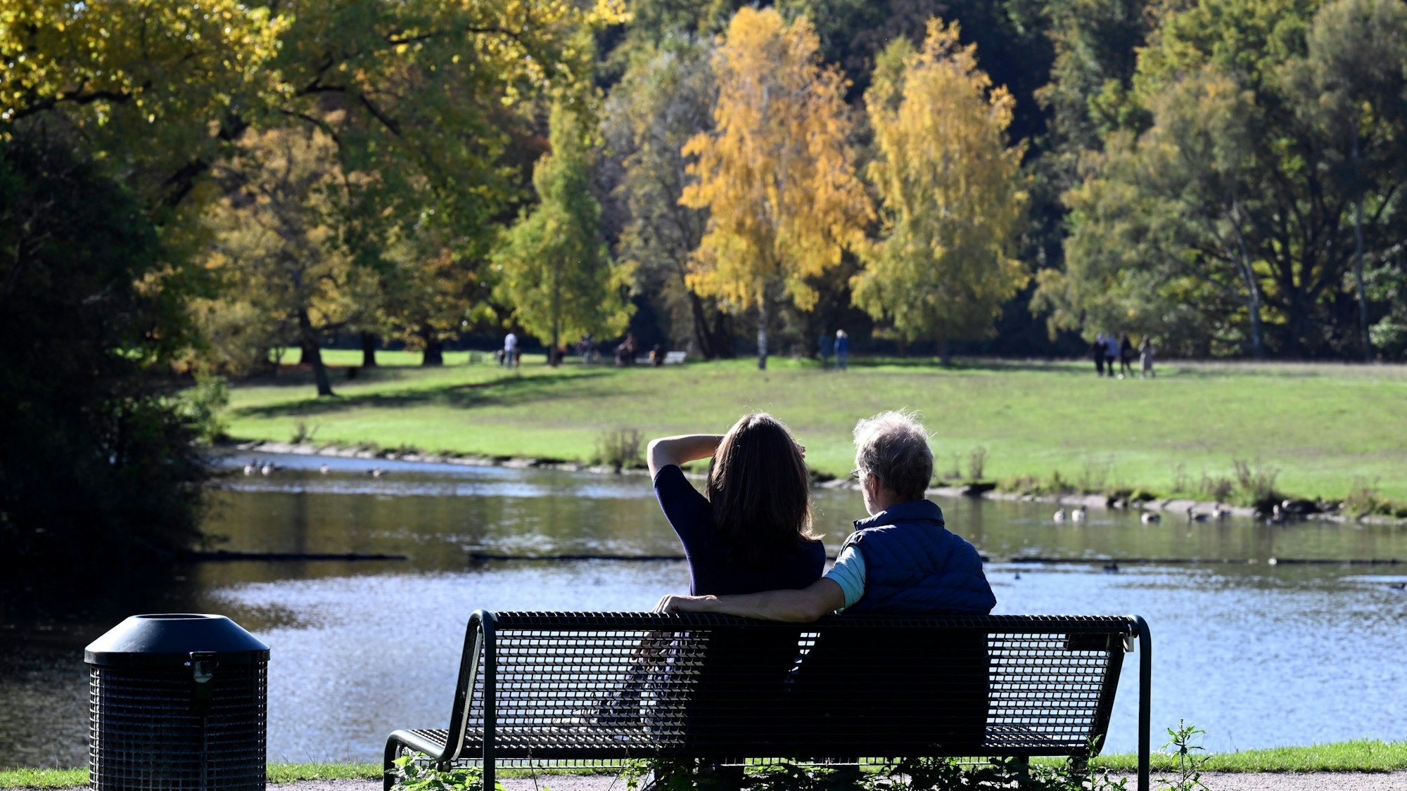 Spaziergänger nutzen den Sonnenschein für eine Pause auf einer Parkbank im Kölner Grüngürtel am Decksteiner Weiher.
